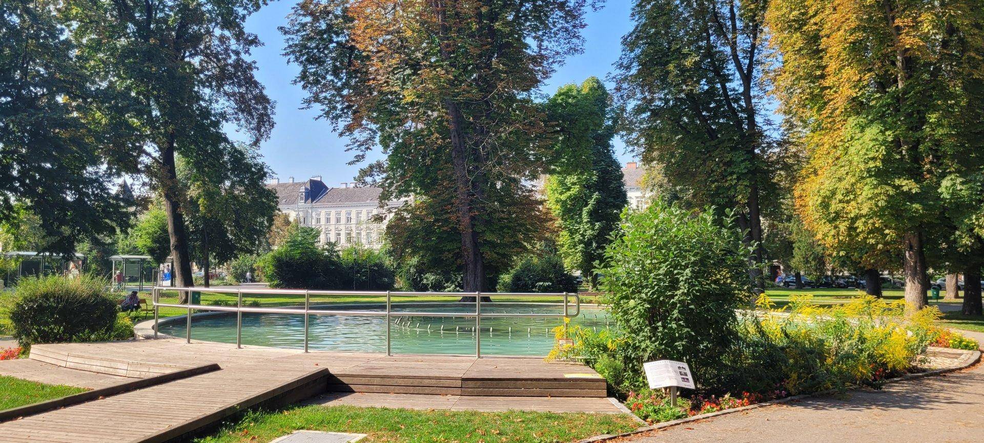 Parkanlage mit einem dekorativen Brunnen, Holzstegen und üppiger Bepflanzung.