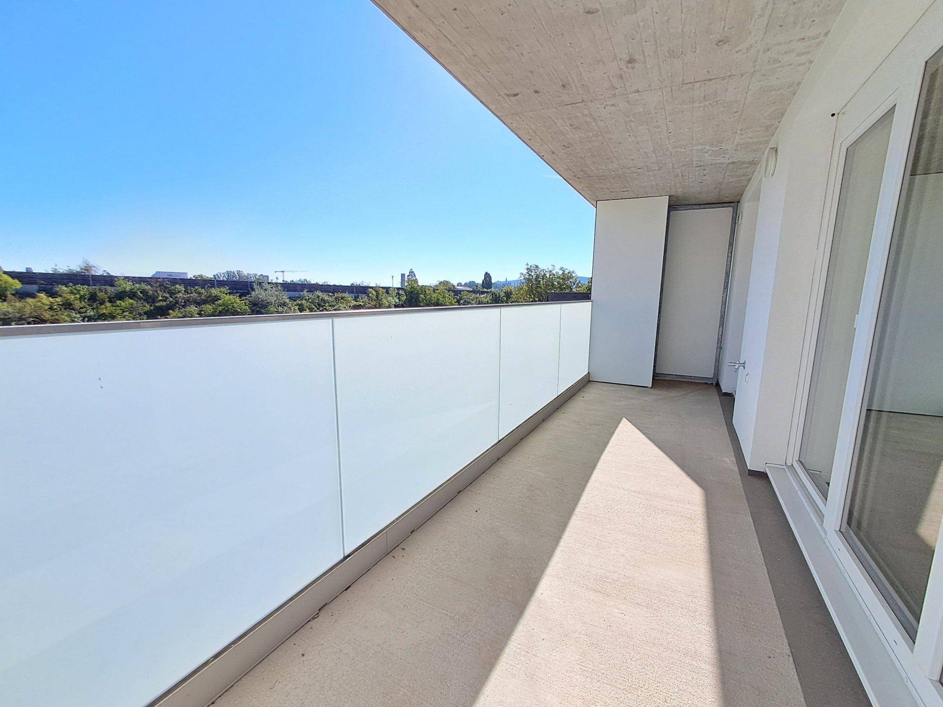 Heller Balkon mit Glasbrüstung und weitem Blick ins Grüne unter blauem Himmel.