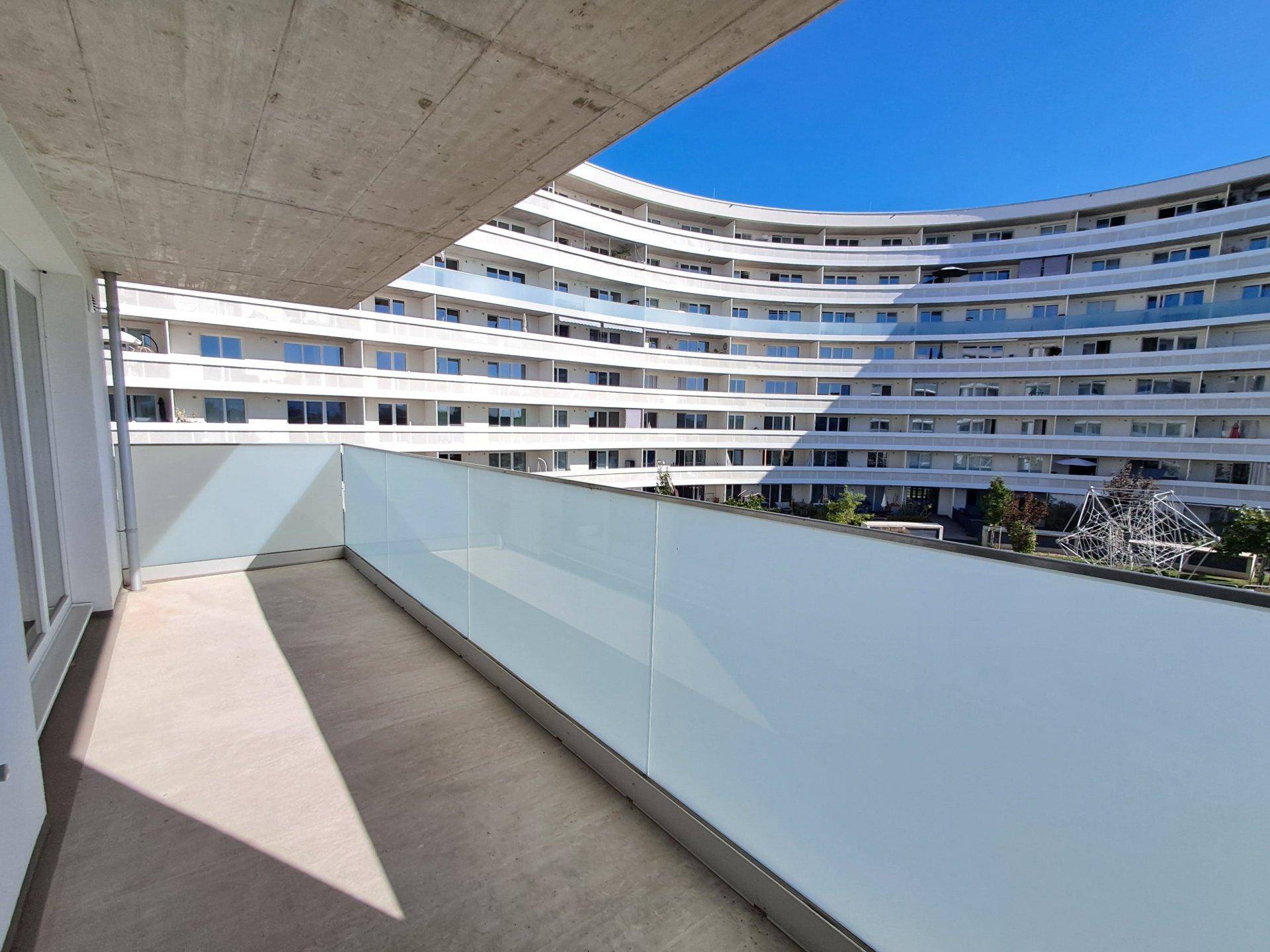 Geräumiger Balkon mit Glasbrüstung und Blick auf ein geschwungenes Nachbargebäude.
