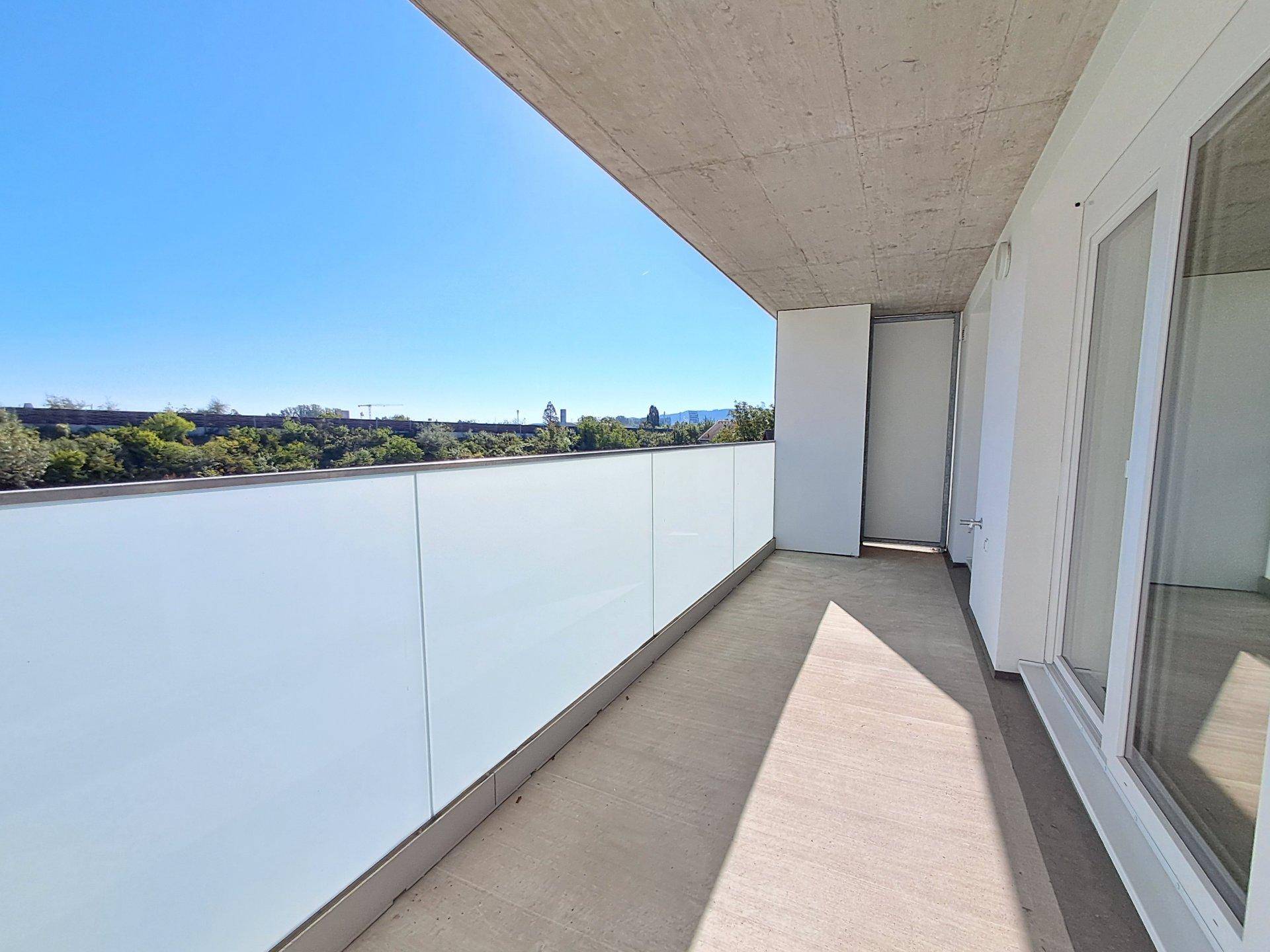 Heller Balkon mit Glasbrüstung und Blick ins Grüne unter blauem Himmel.