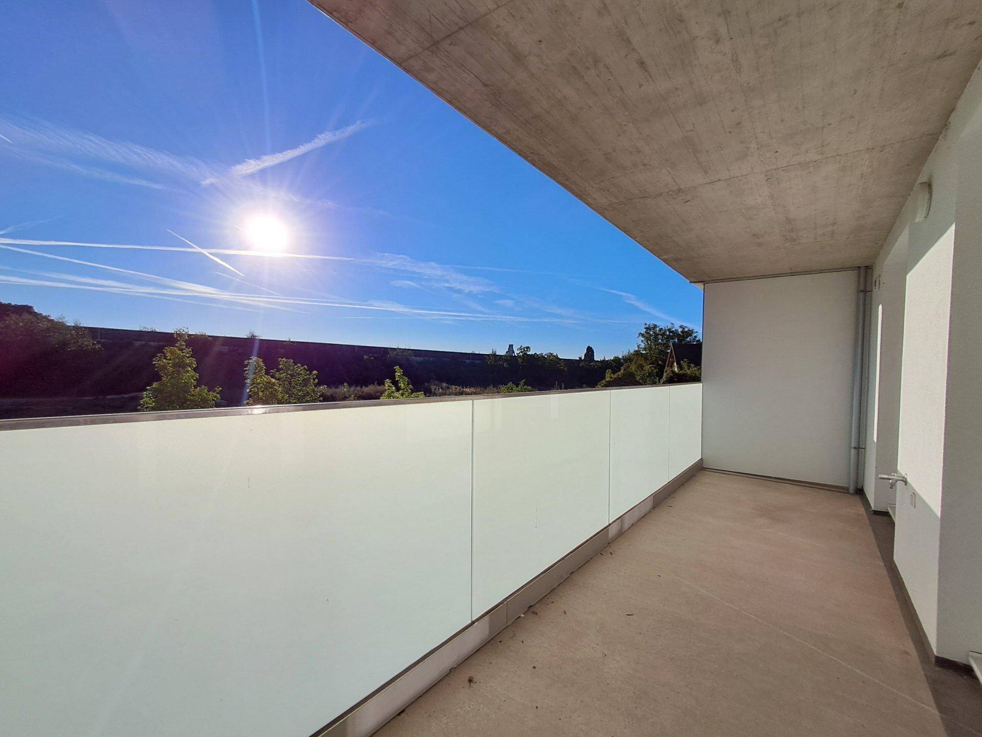 Sonniger Balkon mit moderner Glasbrüstung und weitem Blick ins Grüne unter blauem Himmel.