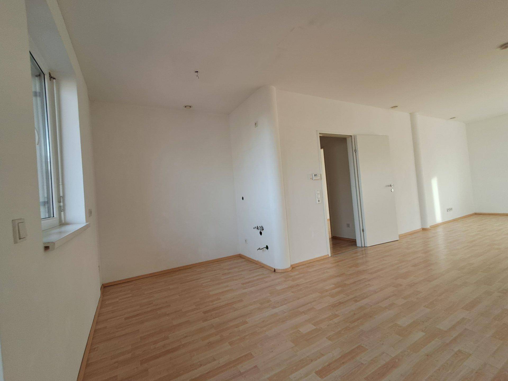 Leerer Raum mit Laminatboden, weißen Wänden und Anschlüssen für eine mögliche Küche.