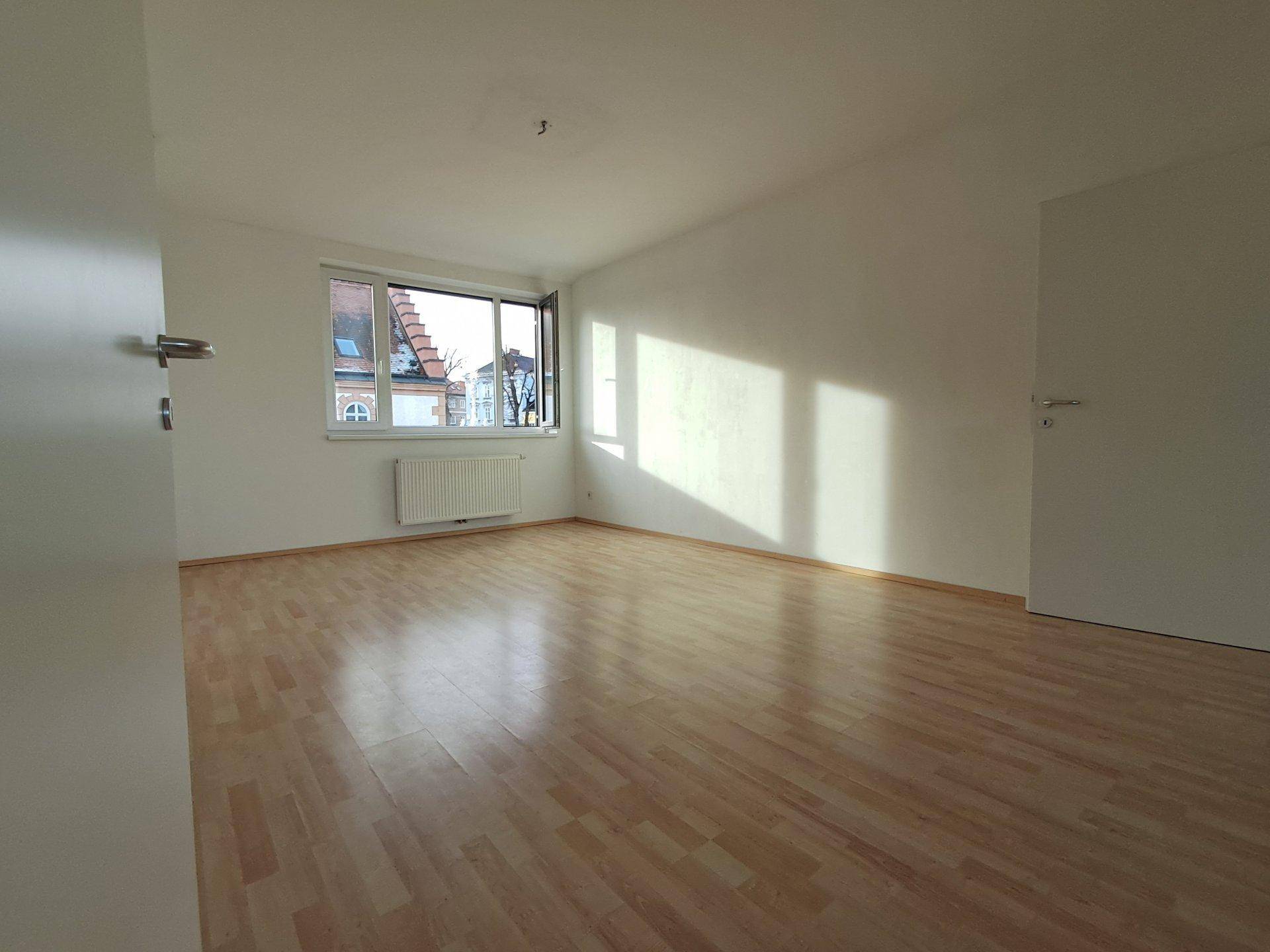 Geräumiger, heller Raum mit Laminatboden und großen Fenstern, ideal als Wohnzimmer.