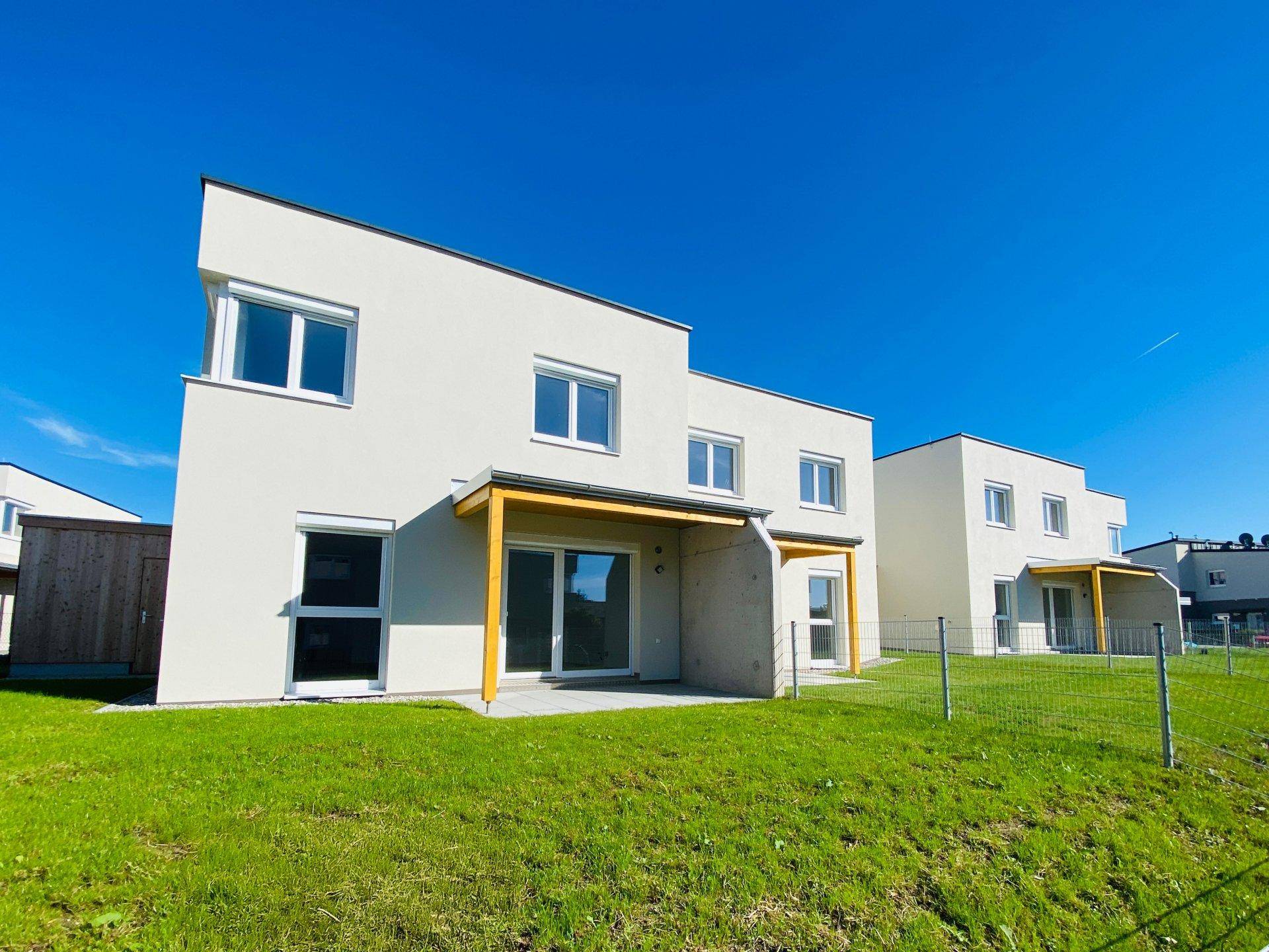 Rückseite der modernen Häuser mit Terrassen und weitläufigen Rasenflächen unter blauem Himmel.