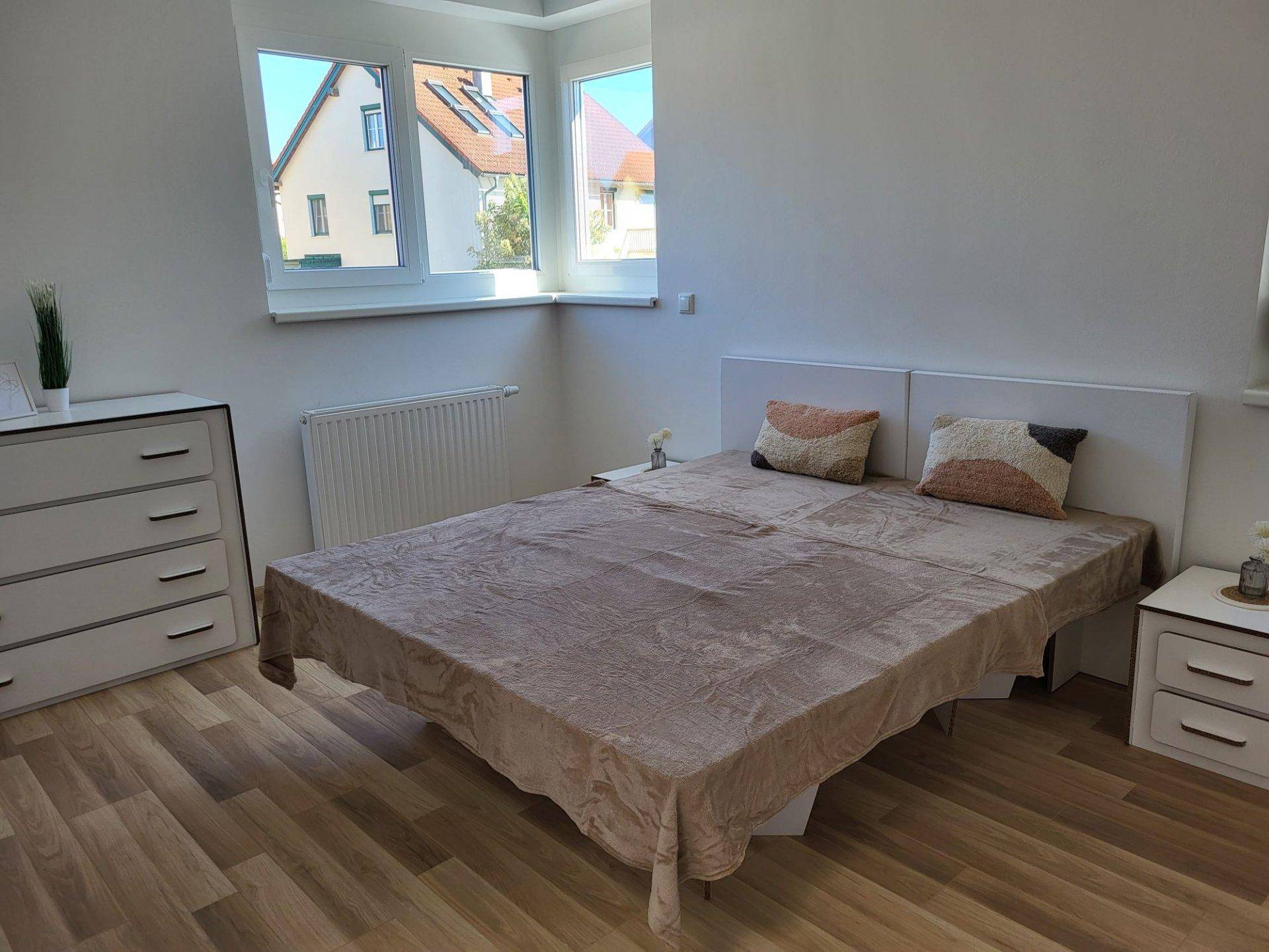 Helles Schlafzimmer mit Laminatboden, großem Fenster und moderner, schlichter Einrichtung.