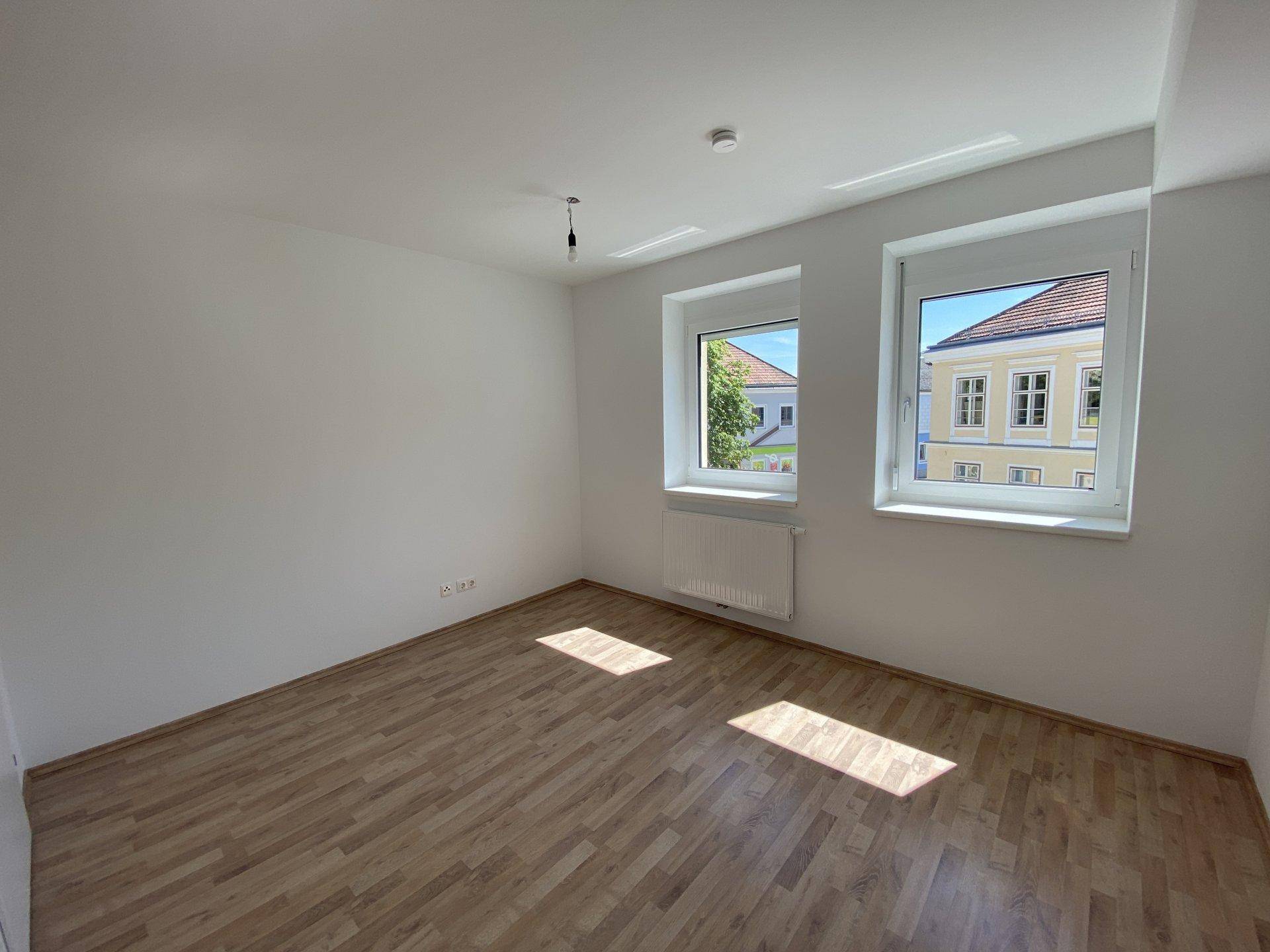 Helles Schlafzimmer mit Laminatboden und zwei Fenstern, die viel Tageslicht hereinlassen.