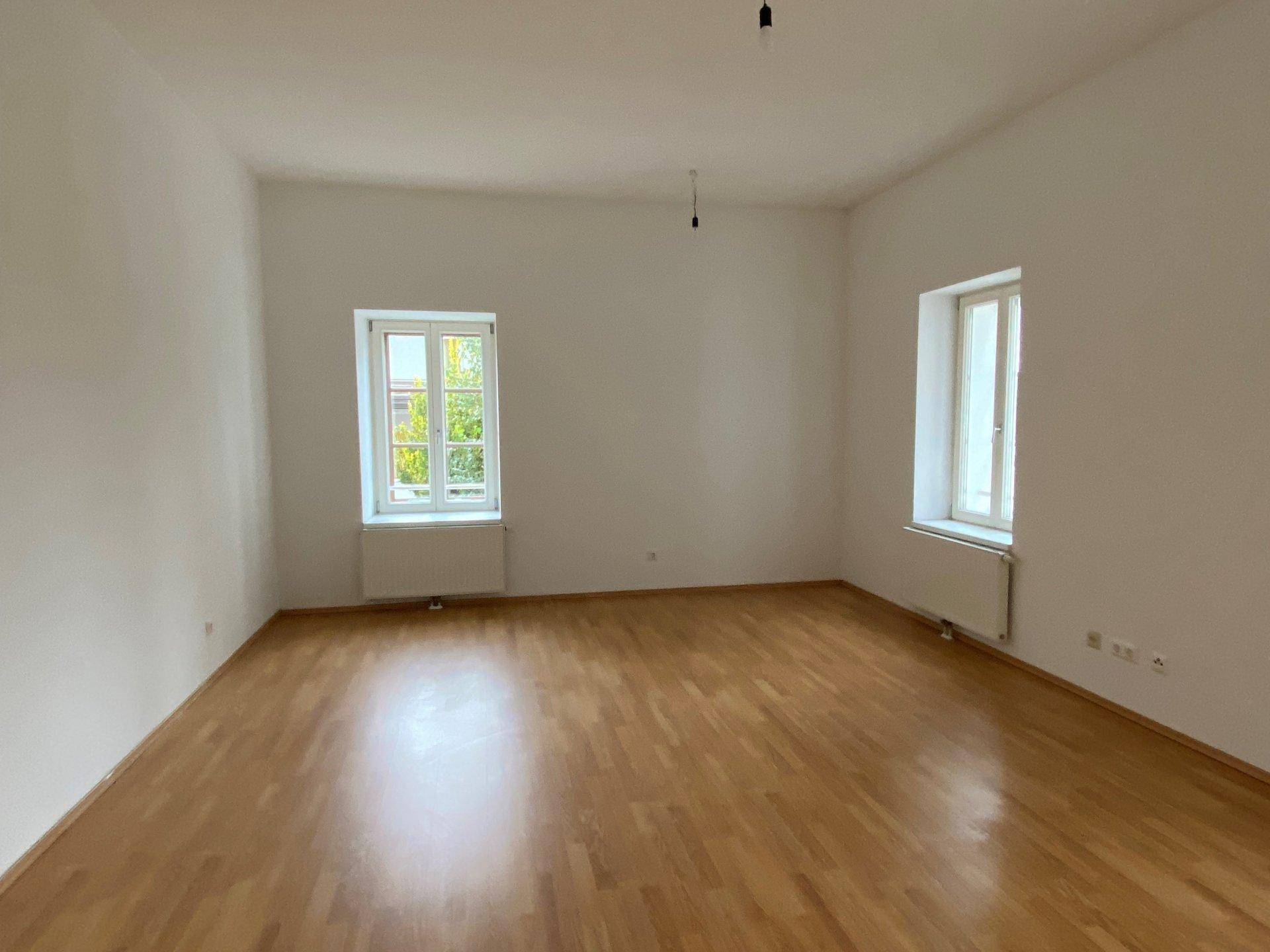 Helles, leeres Zimmer mit Parkettboden und zwei Fenstern, bereit zur individuellen Gestaltung.