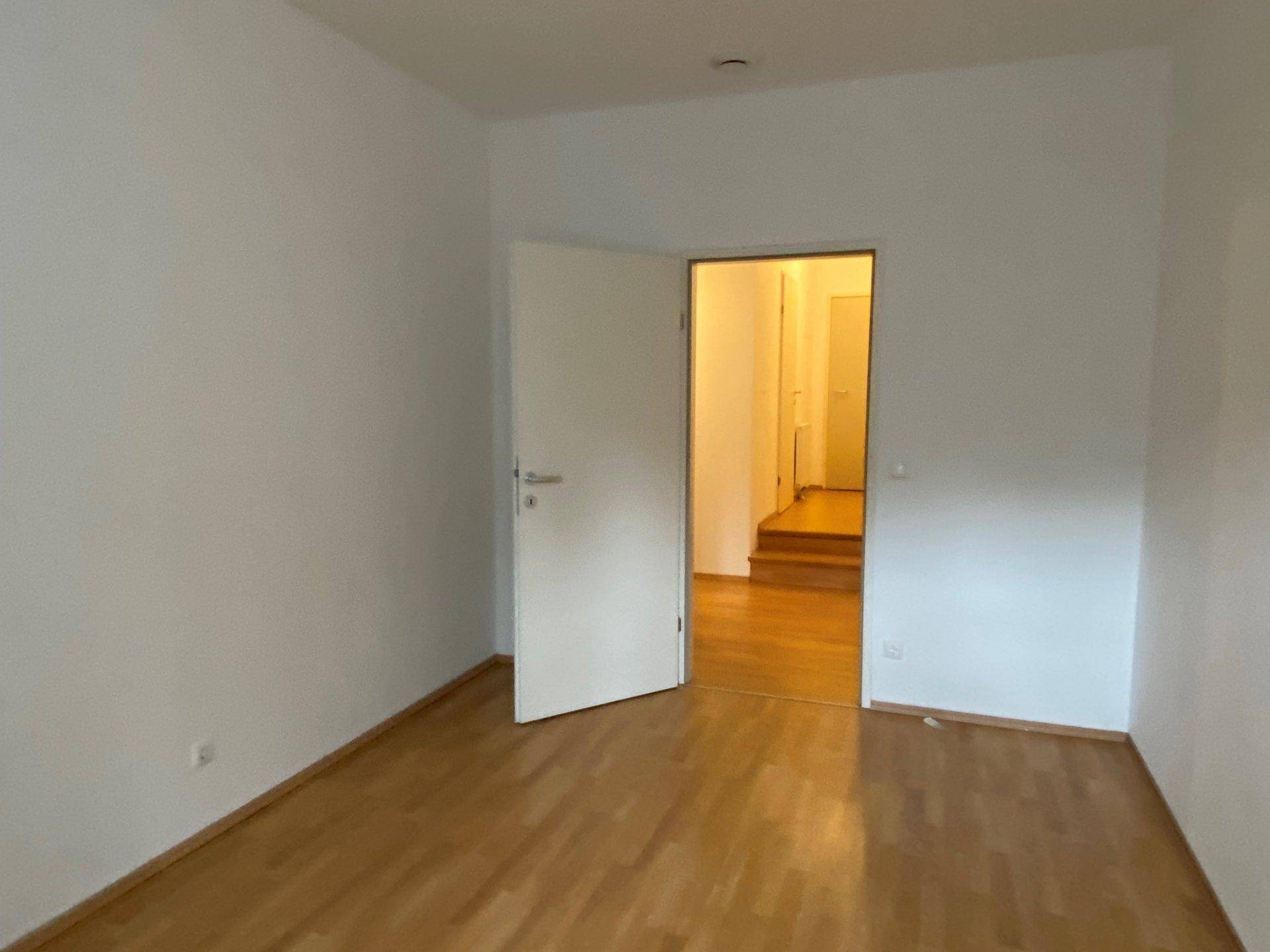 Geräumiges Zimmer mit Holzfußboden und einer Tür, die zu einem Flur mit Treppe führt.