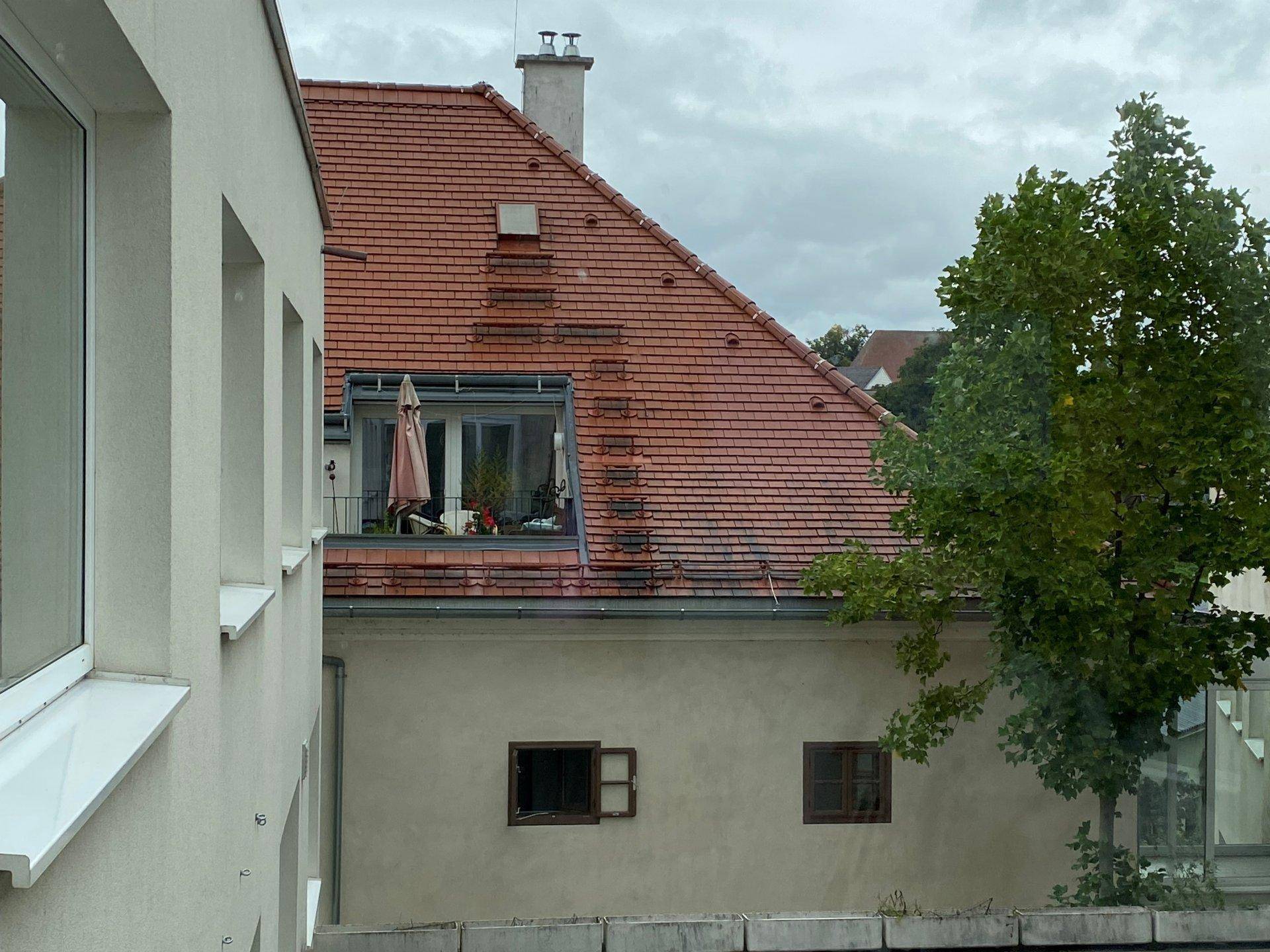 Blick aus dem Fenster auf ein benachbartes Dach mit Dachziegeln und einem kleinen Balkon.