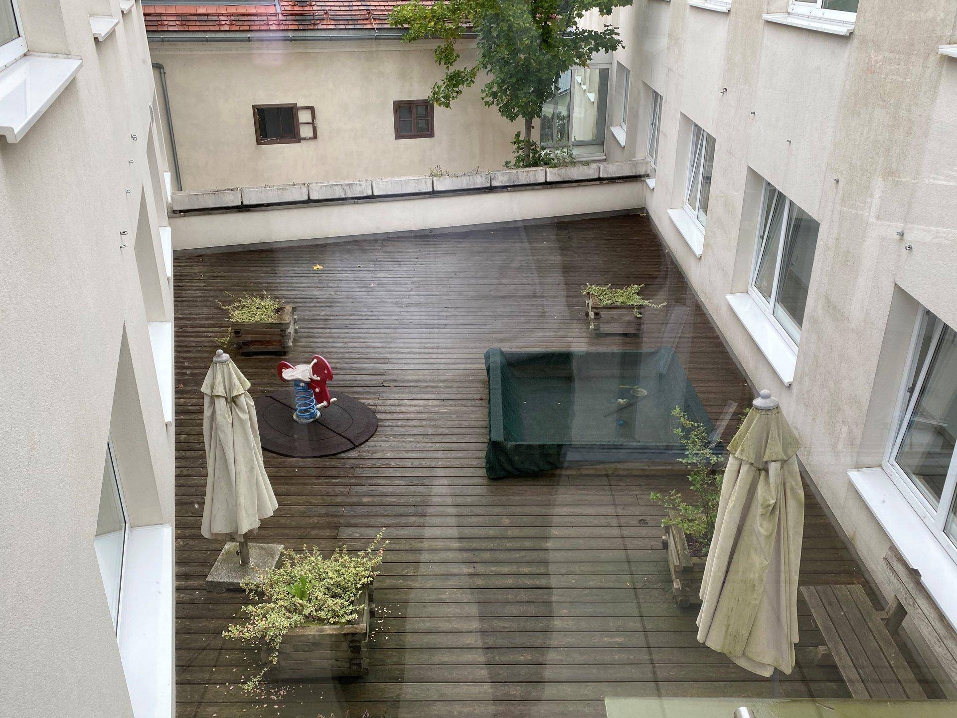 Blick von oben auf einen Innenhof mit Holzterrasse, Sonnenschirmen und Spielgeräten.
