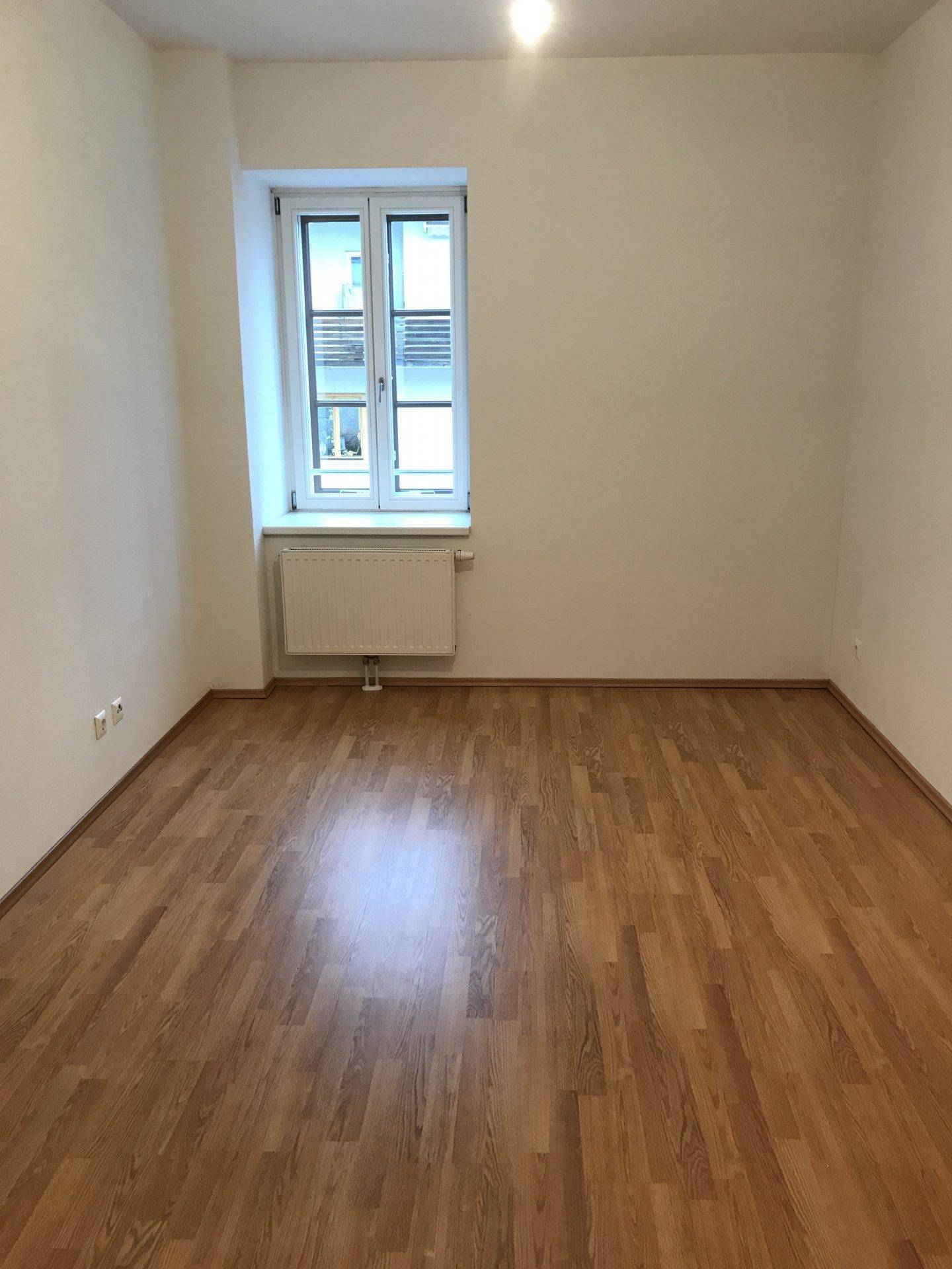Leeres Zimmer mit Holzfußboden und einem Fenster, das viel Tageslicht hereinlässt.