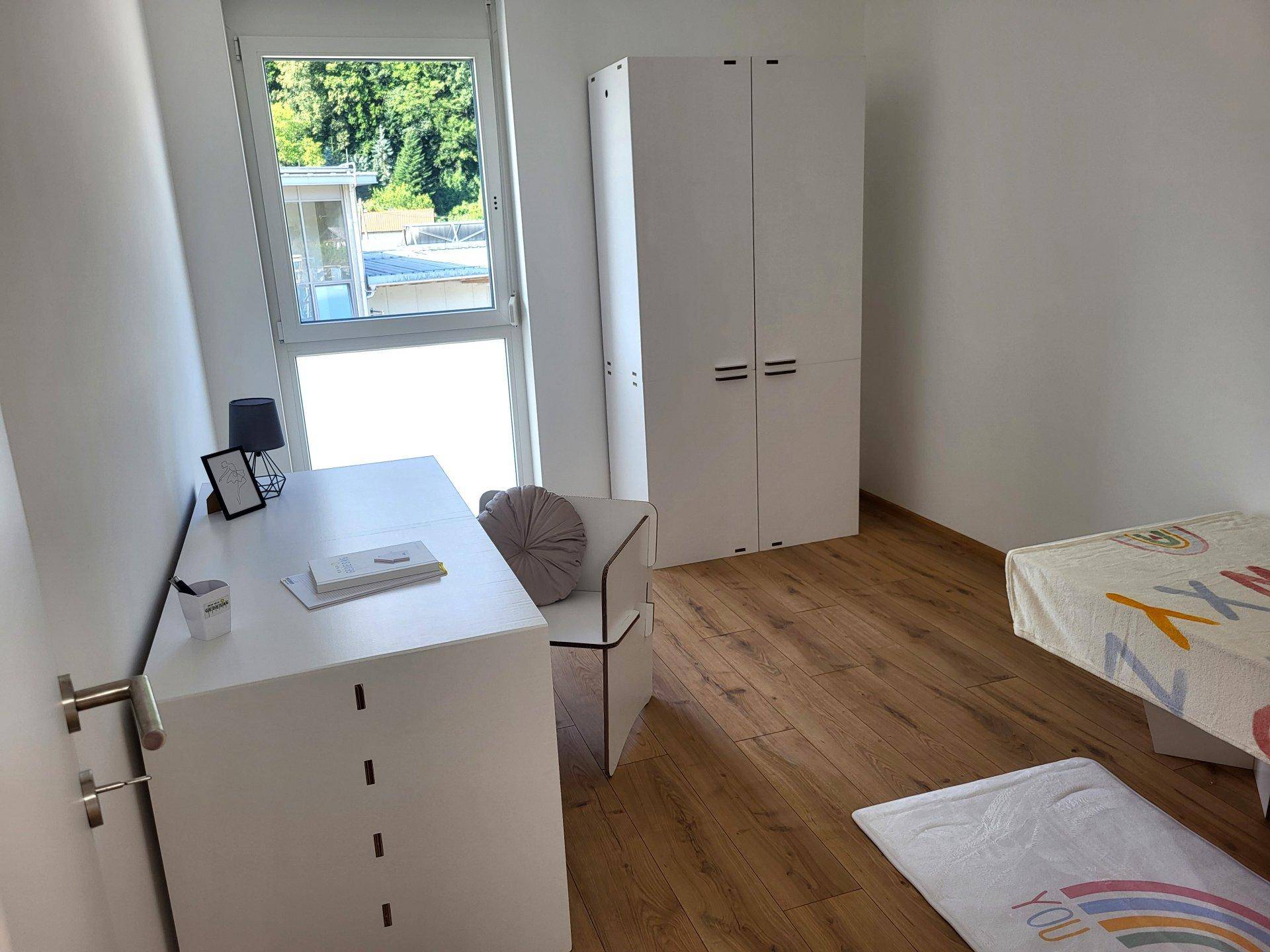 Kinderzimmer mit Schreibtisch, Kleiderschrank und großem Fenster auf Holzboden.