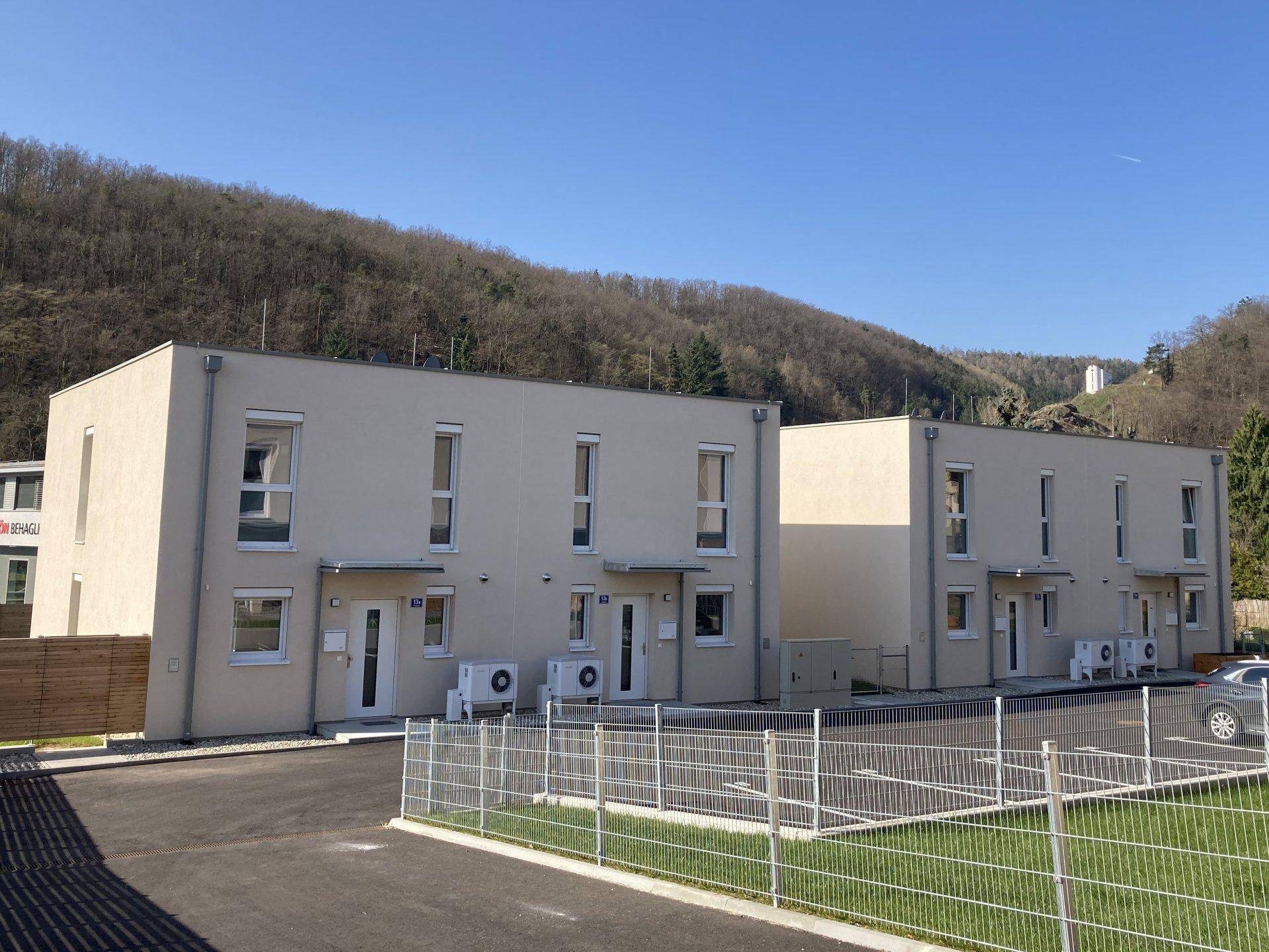 Moderne Reihenhäuser mit heller Fassade, Parkplätzen und Blick auf bewaldete Hügel.