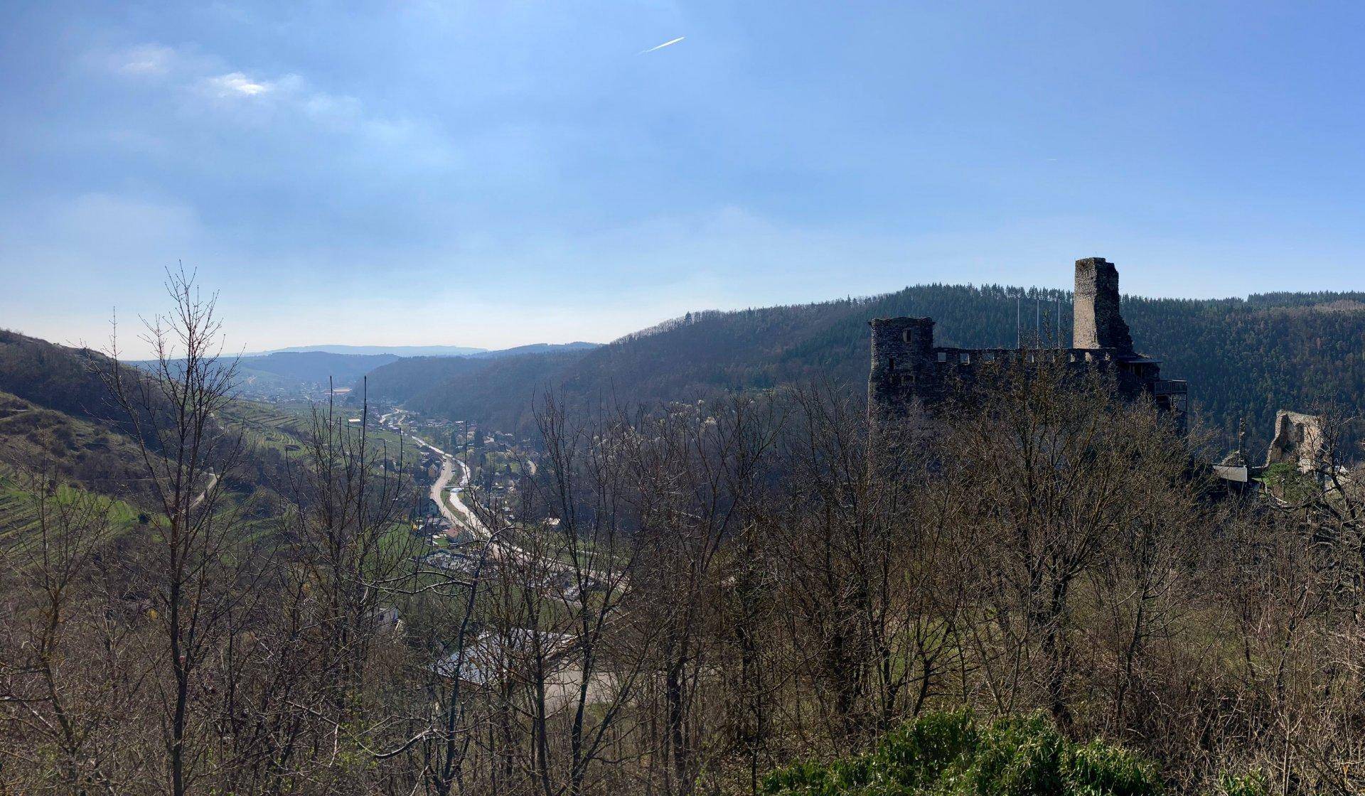 Malerischer Ausblick auf eine Burgruine, ein Tal und bewaldete Berge unter blauem Himmel.