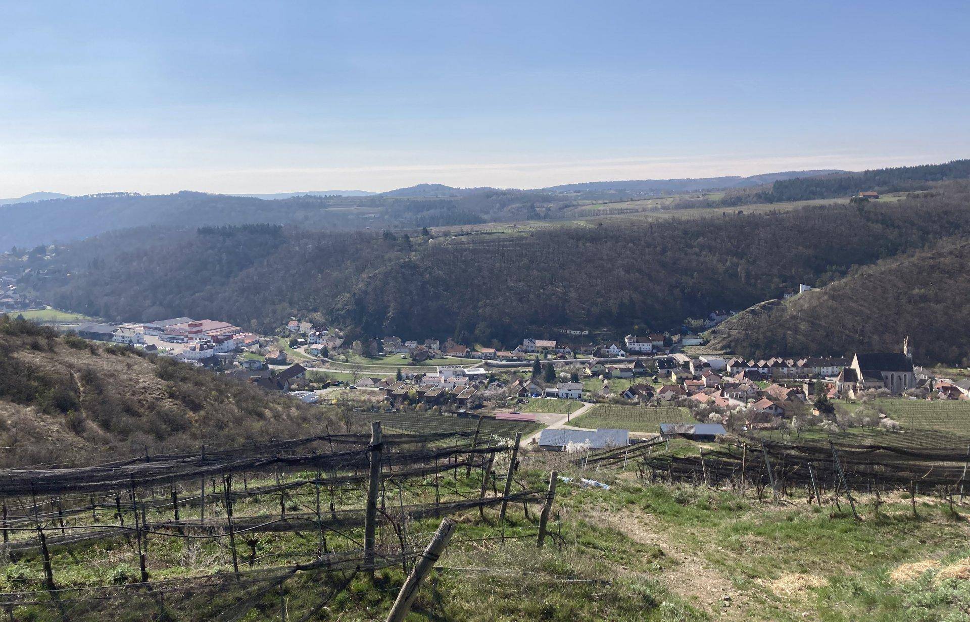 Weitläufiger Blick über ein Tal mit Weinbergen, einem Dorf und bewaldeten Hügeln.