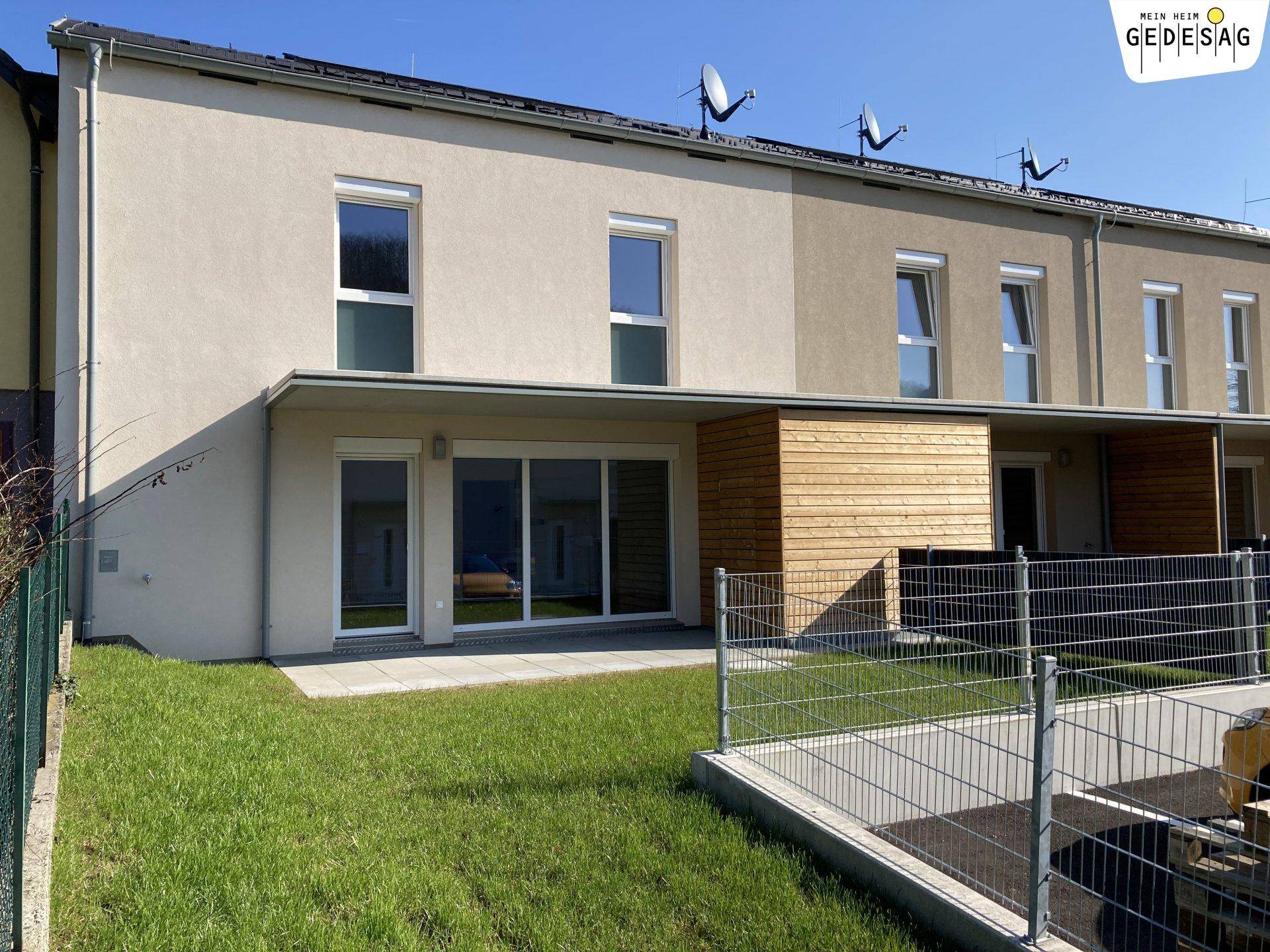 Rückseite eines Reihenhauses mit Terrasse, Rasenfläche und Blick auf die Umgebung.