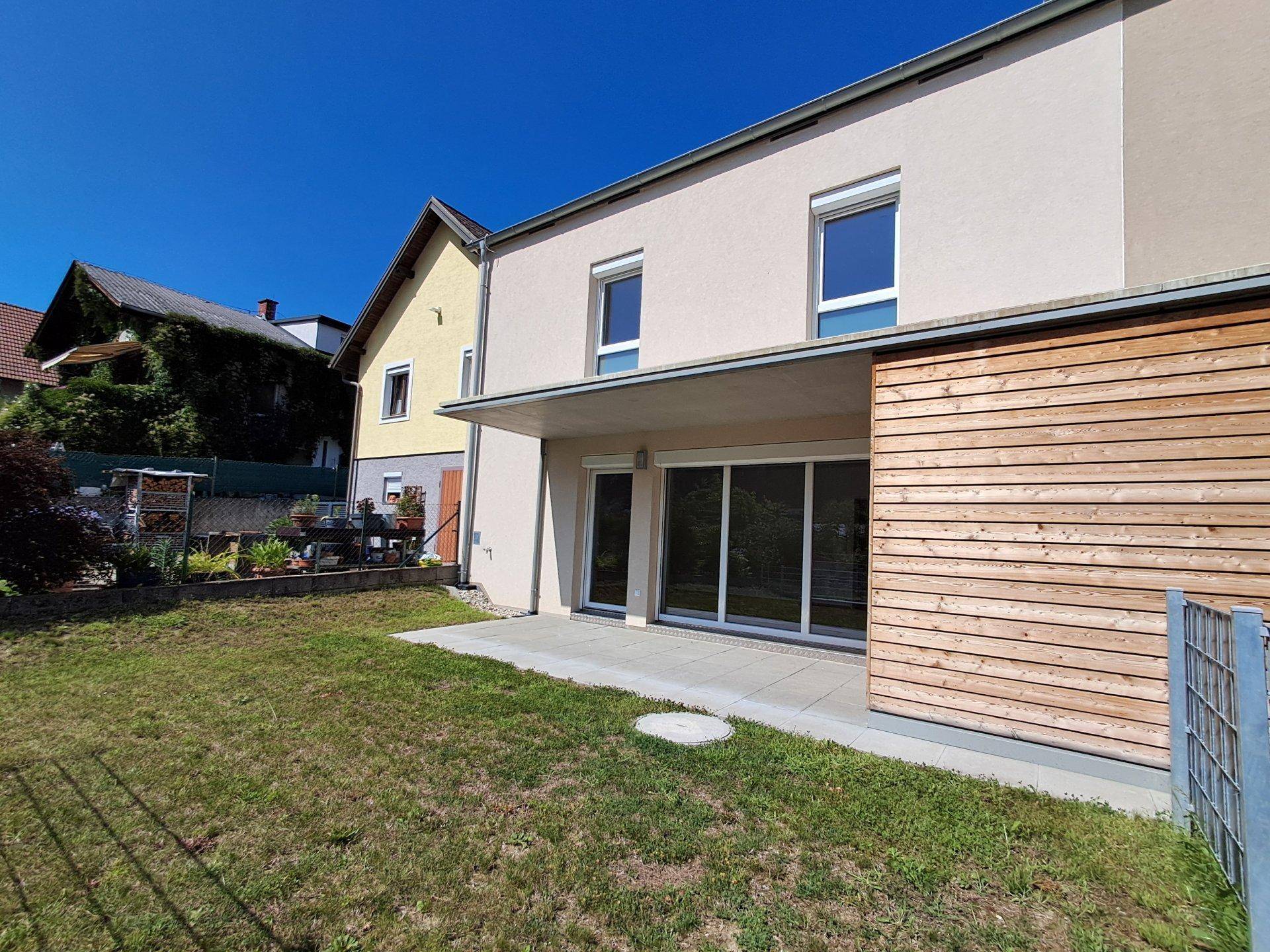 Rückseite eines Reihenhauses mit Terrasse und gepflegtem Gartenbereich unter blauem Himmel.