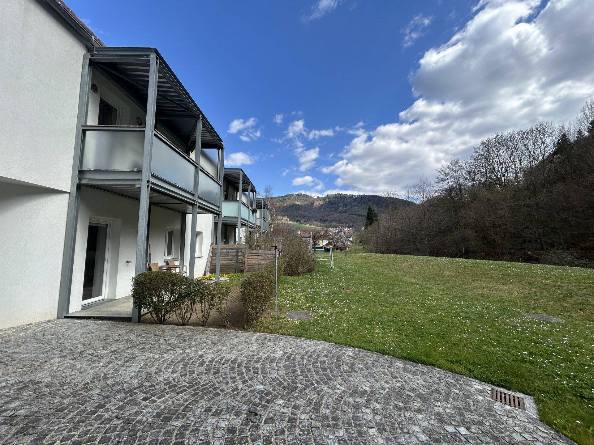 Moderne Wohngebäude mit Balkonen und gepflastertem Vorplatz, umgeben von grünen Wiesen und Hügeln.