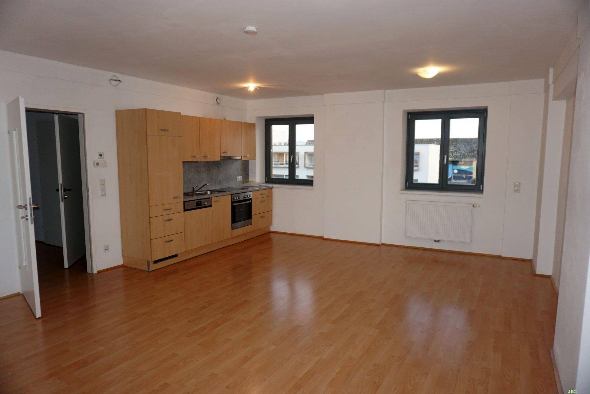 Offener Wohnbereich mit integrierter Küchenzeile, Parkettboden und zwei Fenstern für Tageslicht.