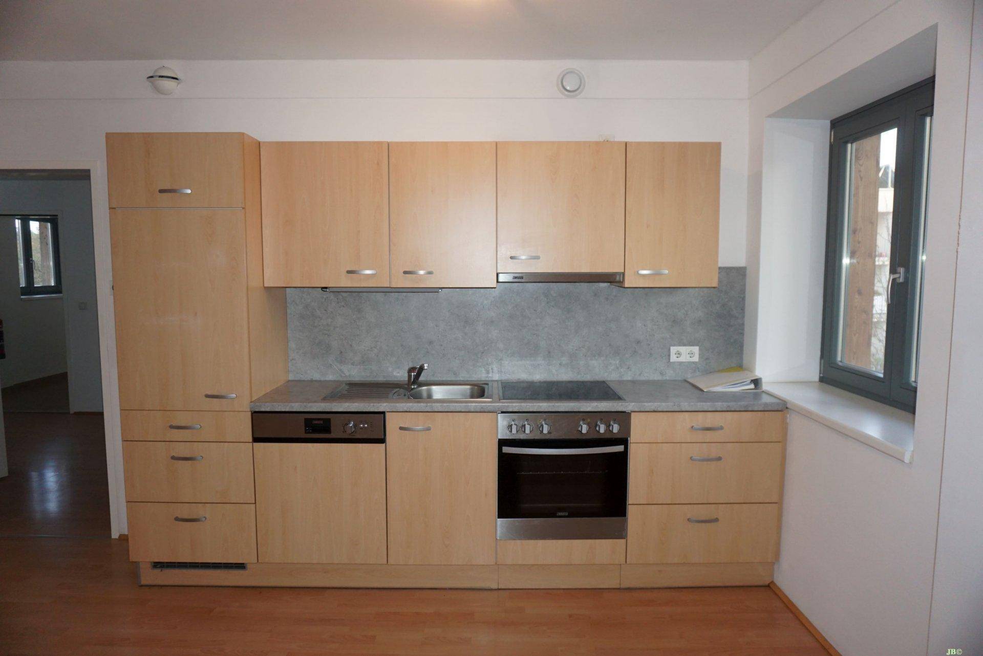 Funktionale Einbauküche mit hellen Holzfronten, Spülmaschine, Backofen und Fenster.