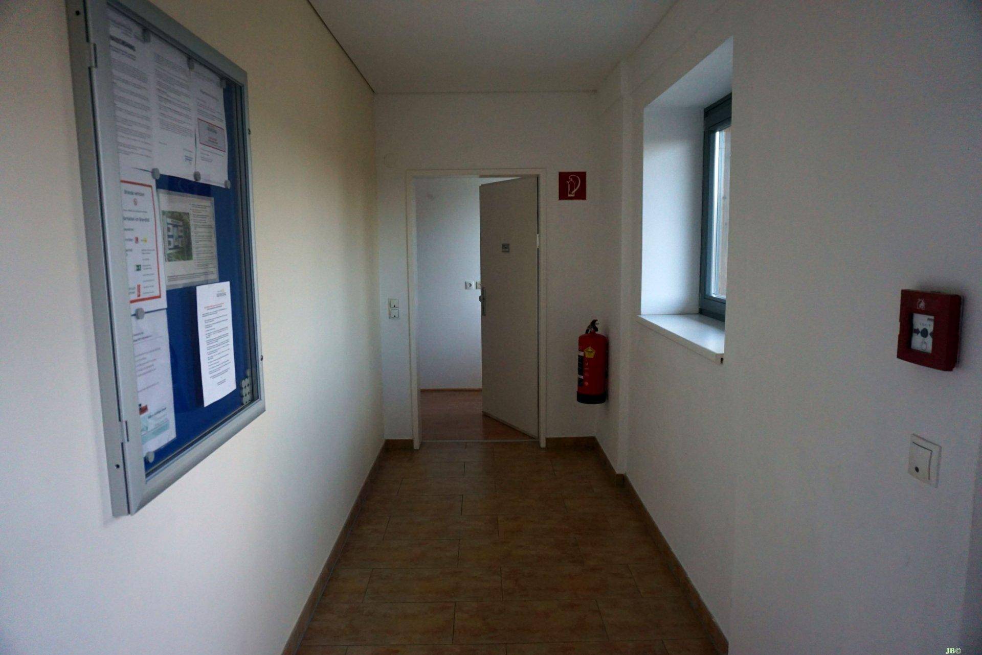 Heller Flur mit Fliesenboden, Informationspinnwand und Zugang zu weiteren Räumen.