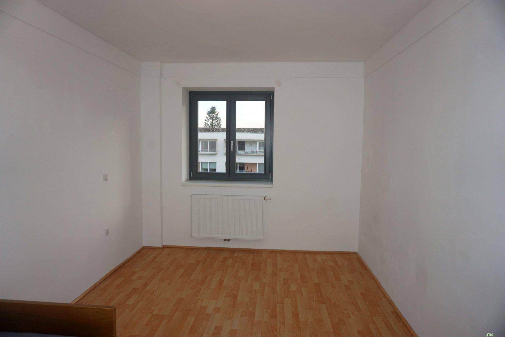 Leeres Schlafzimmer mit hellem Parkettboden und Fenster mit Blick ins Grüne.