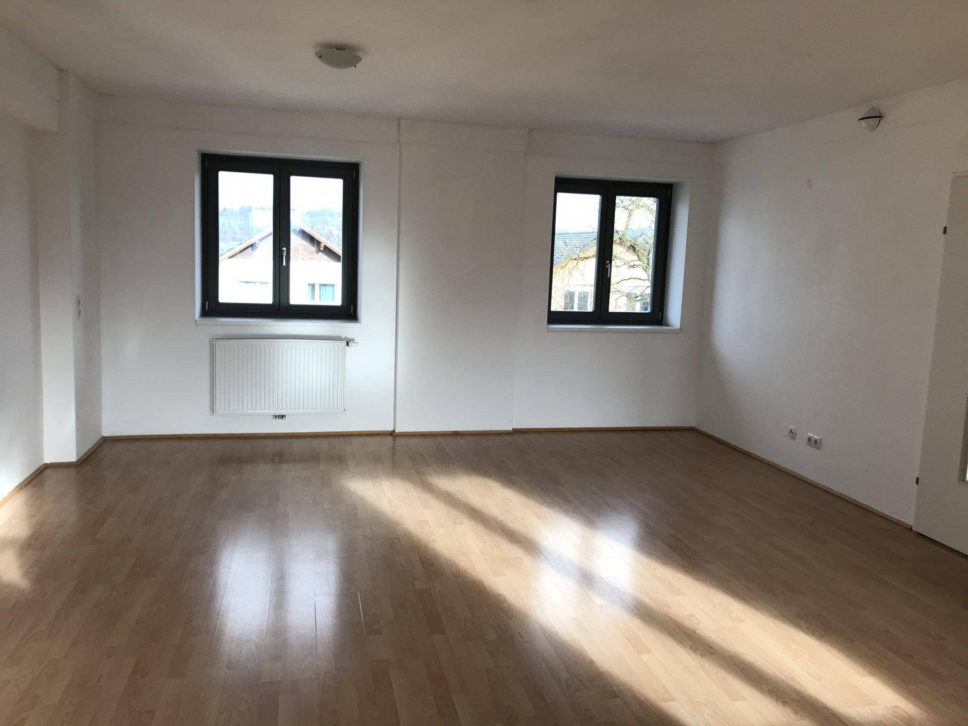 Geräumiges Wohnzimmer mit hellem Parkettboden und zwei Fenstern, die viel Sonnenlicht hereinlassen.