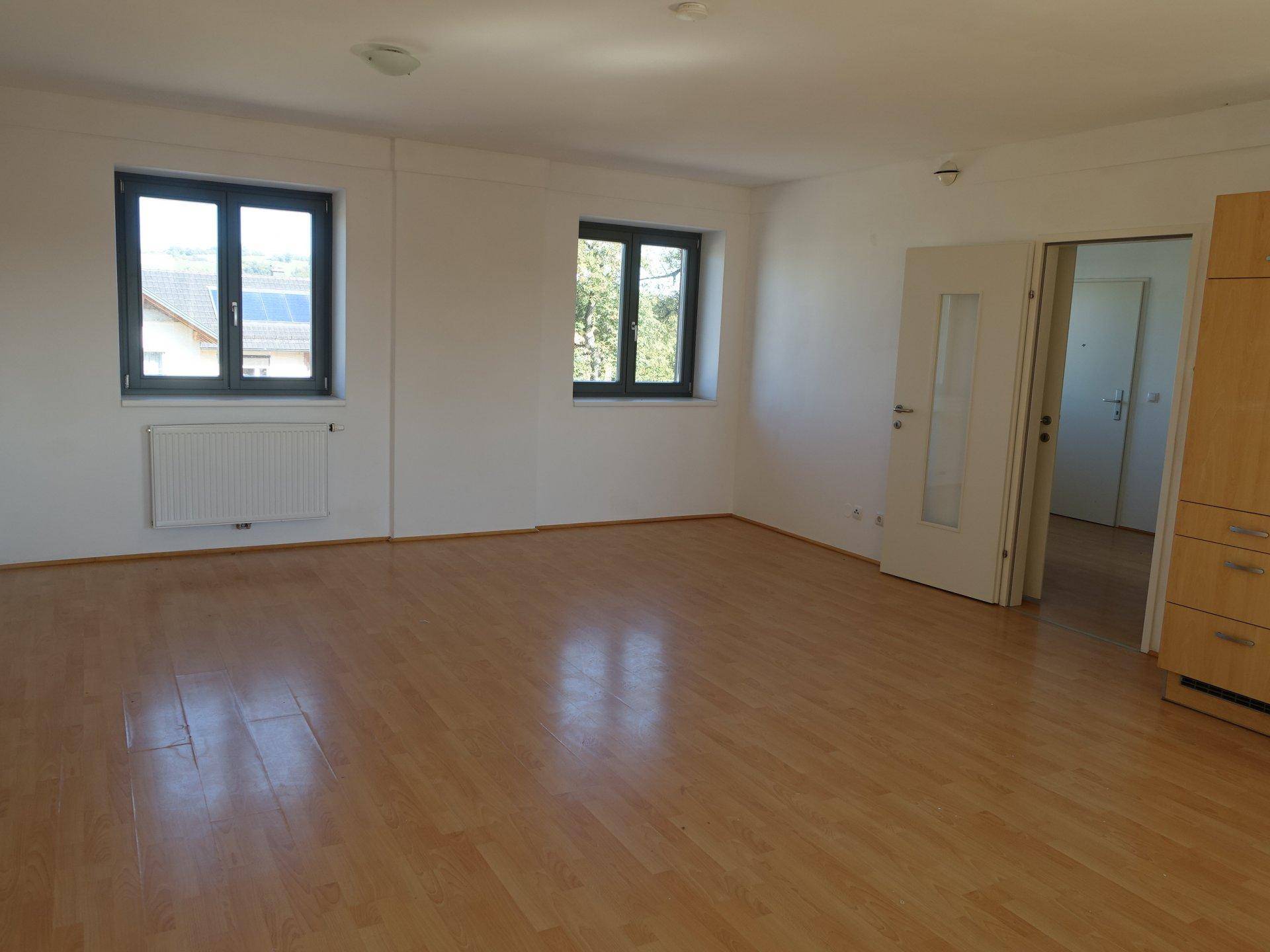 Geräumiger Wohnbereich mit hellem Parkettboden und zwei Fenstern, die viel Tageslicht bieten.