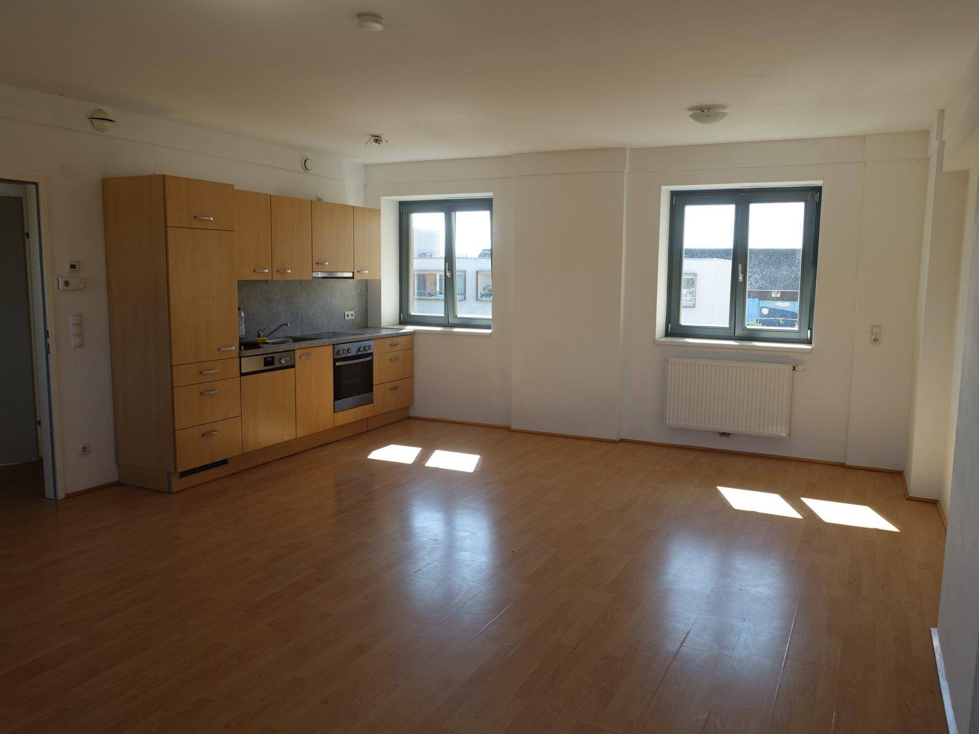 Lichtdurchfluteter Wohnbereich mit integrierter Küchenzeile, Parkettboden und zwei Fenstern.