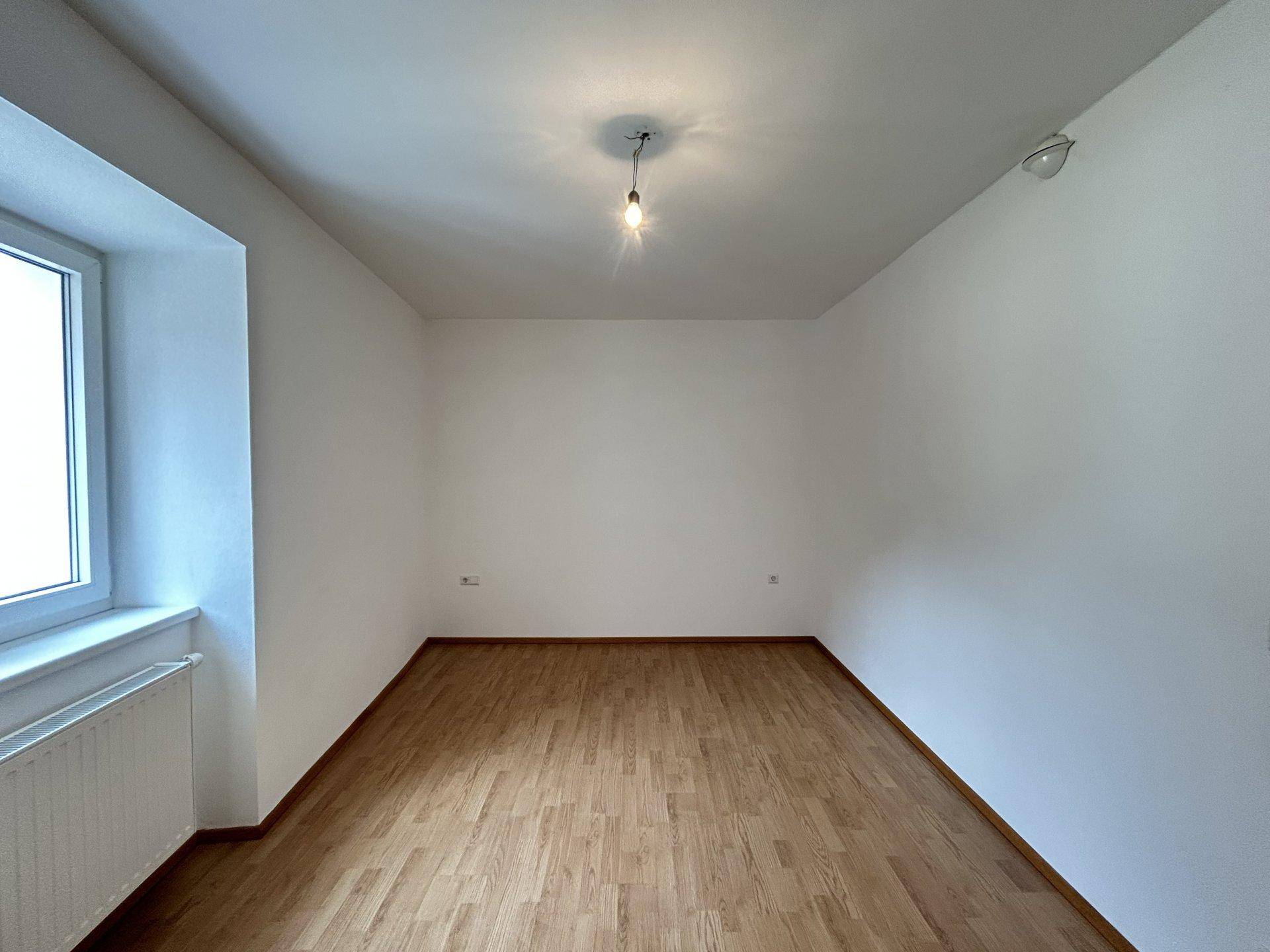 Leeres Schlafzimmer mit Holzboden, weißen Wänden und einem Fenster, das viel Tageslicht bietet.