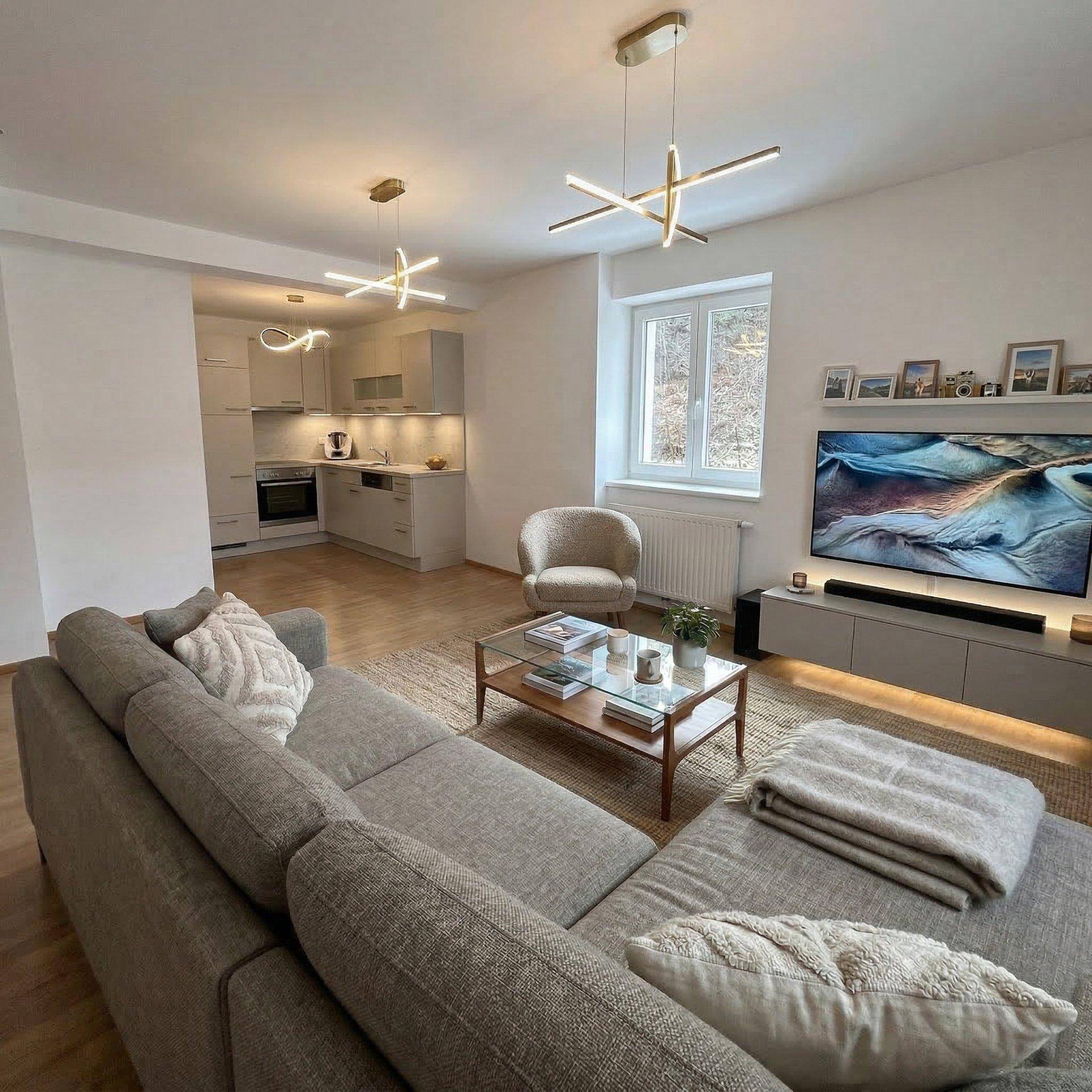 Modernes Wohnzimmer mit offener Küche, bequemer Couch, Fernseher und stilvoller Beleuchtung.