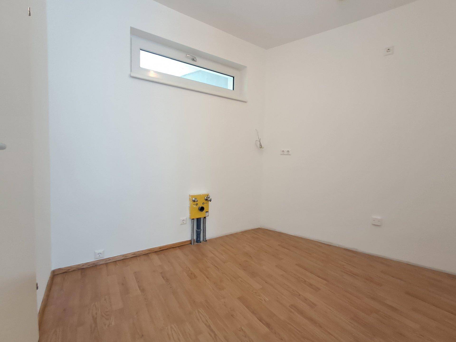 Kleiner Raum mit Laminatboden, Fenster und Anschlüssen für Sanitärinstallationen.