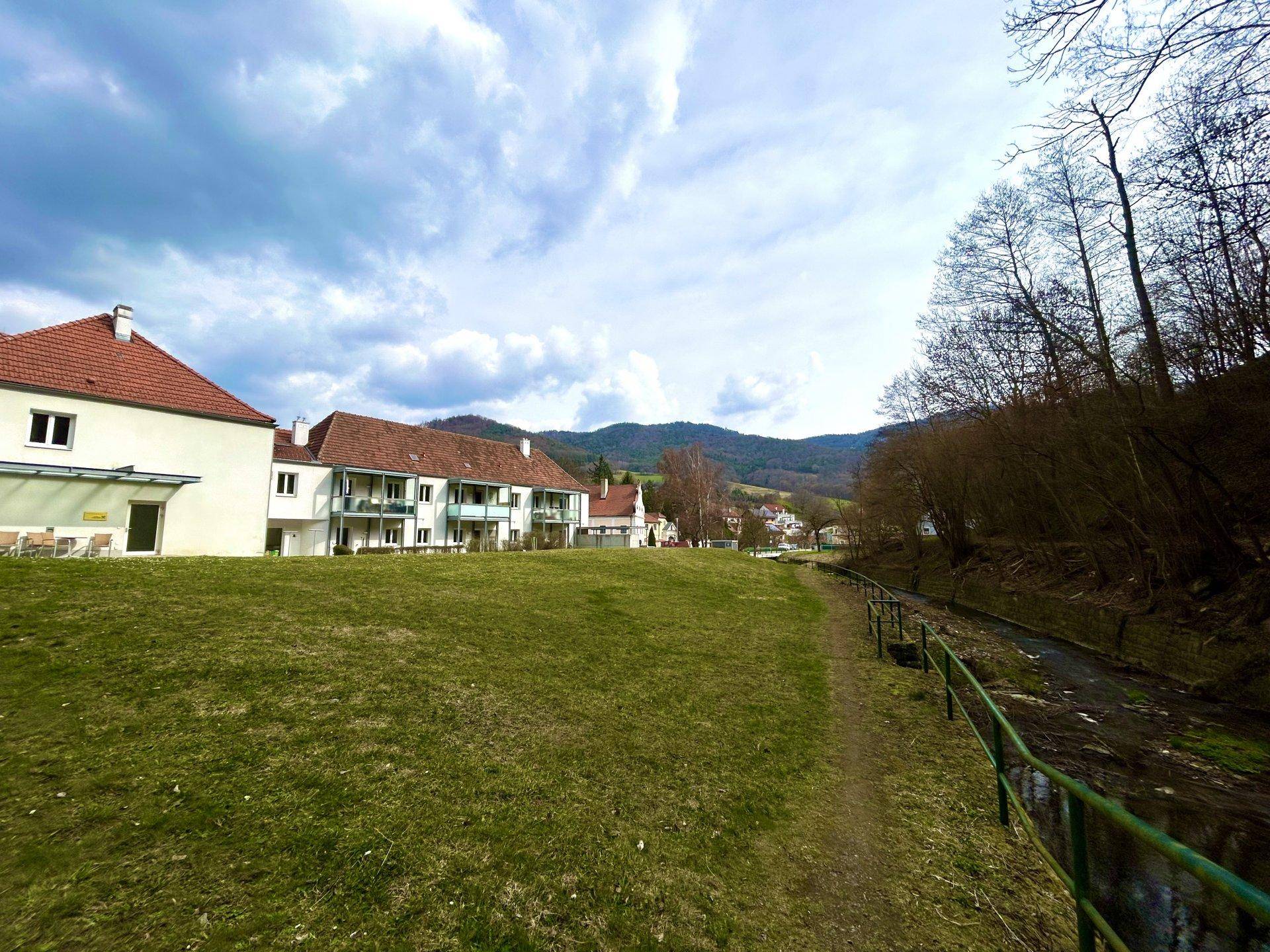 Blick auf Wohngebäude mit roten Dächern, eine weite Rasenfläche und einen Bachlauf am Waldrand.