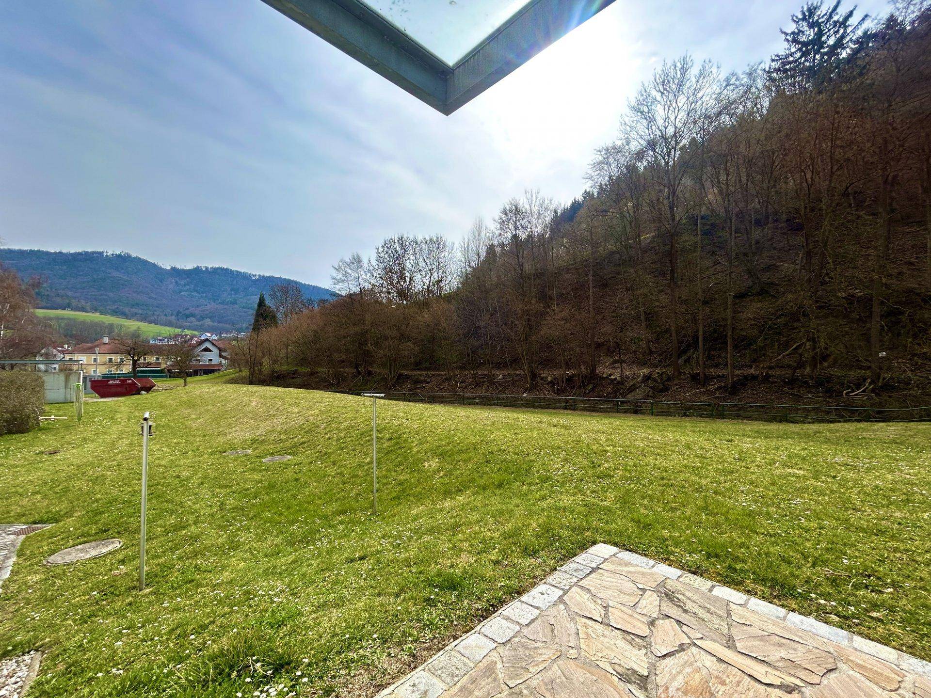 Große, sonnige Rasenfläche mit einer Steinterrasse und Blick auf bewaldete Hügel und Natur.