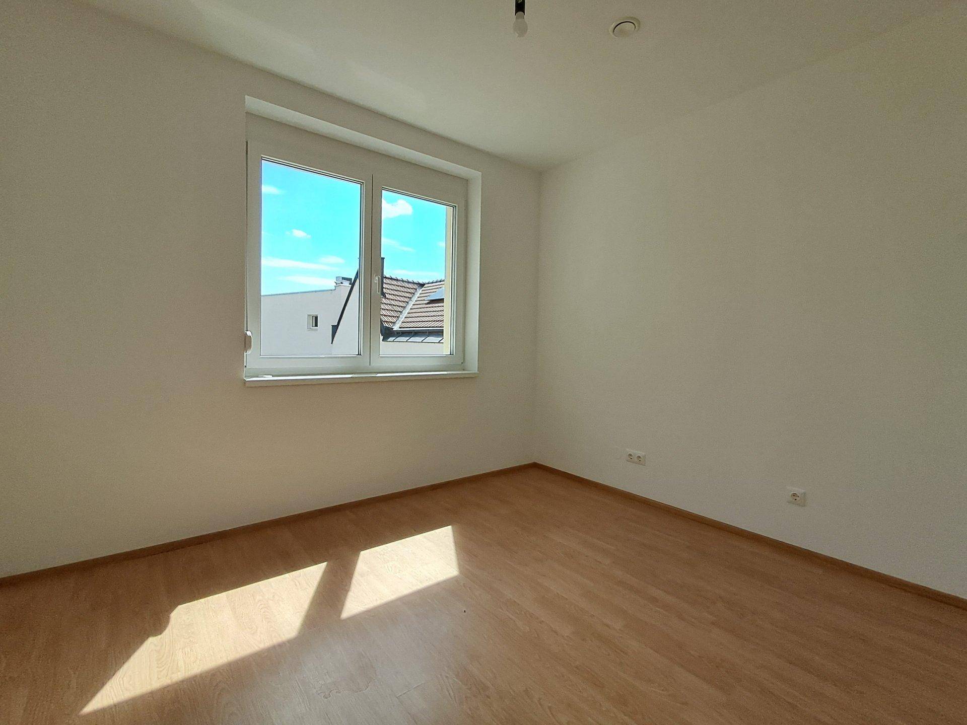 Helles Schlafzimmer mit Laminatboden und großem Fenster, das viel Sonnenlicht hereinlässt.
