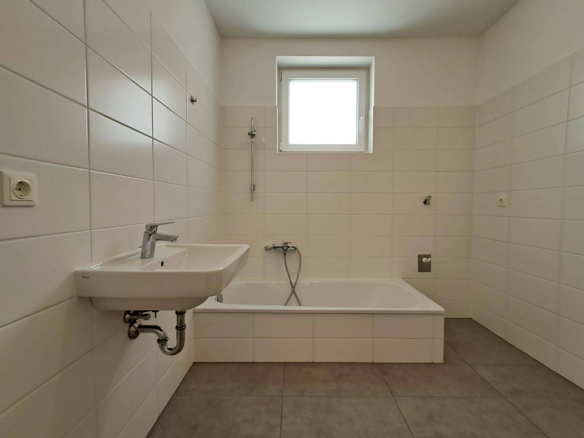 Helles Badezimmer mit Badewanne, Waschbecken und Fenster, ausgestattet mit weißen Fliesen.