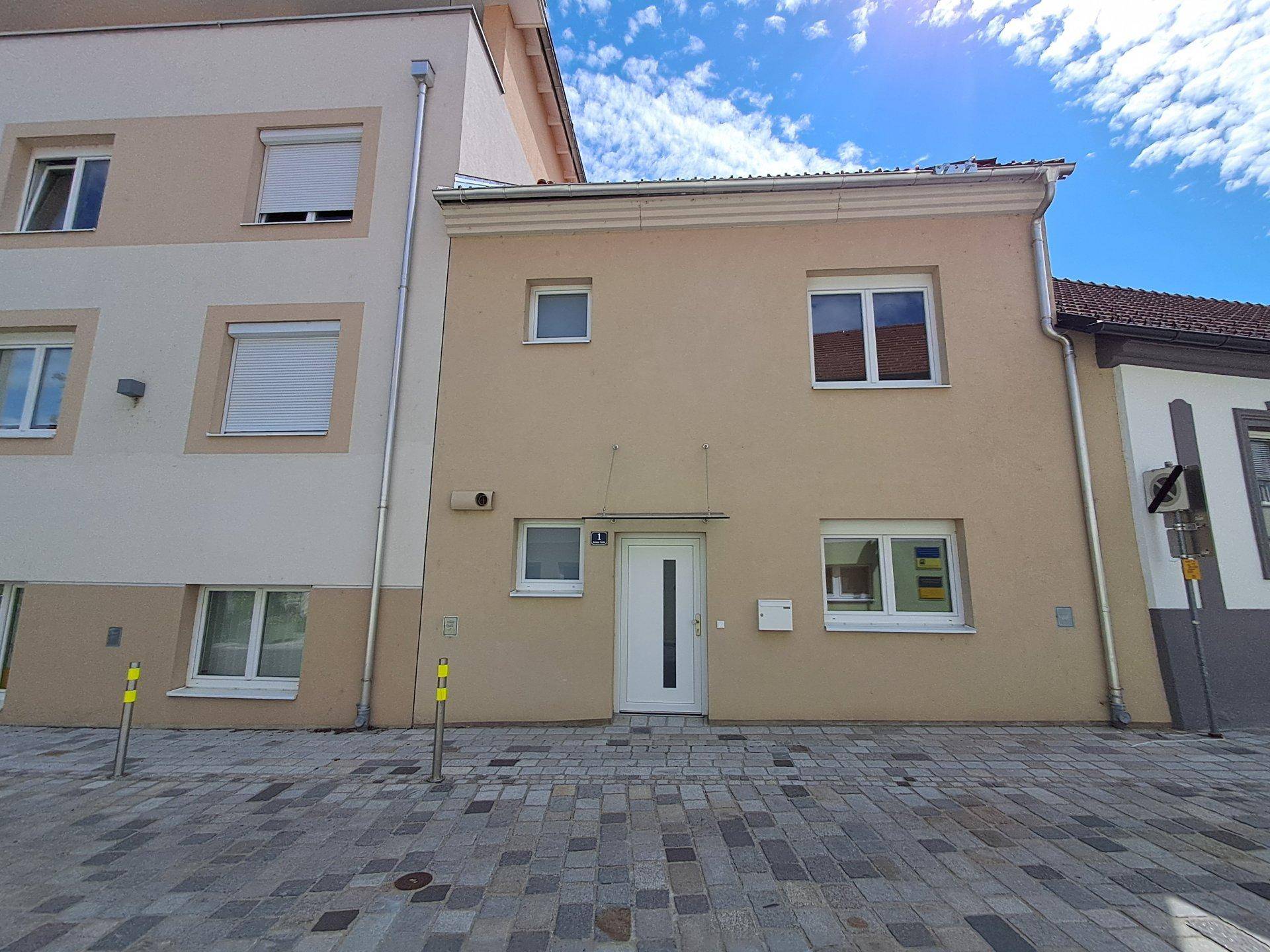 Eingangsbereich eines modernen Gebäudes mit heller Fassade und gepflastertem Vorplatz unter blauem Himmel.
