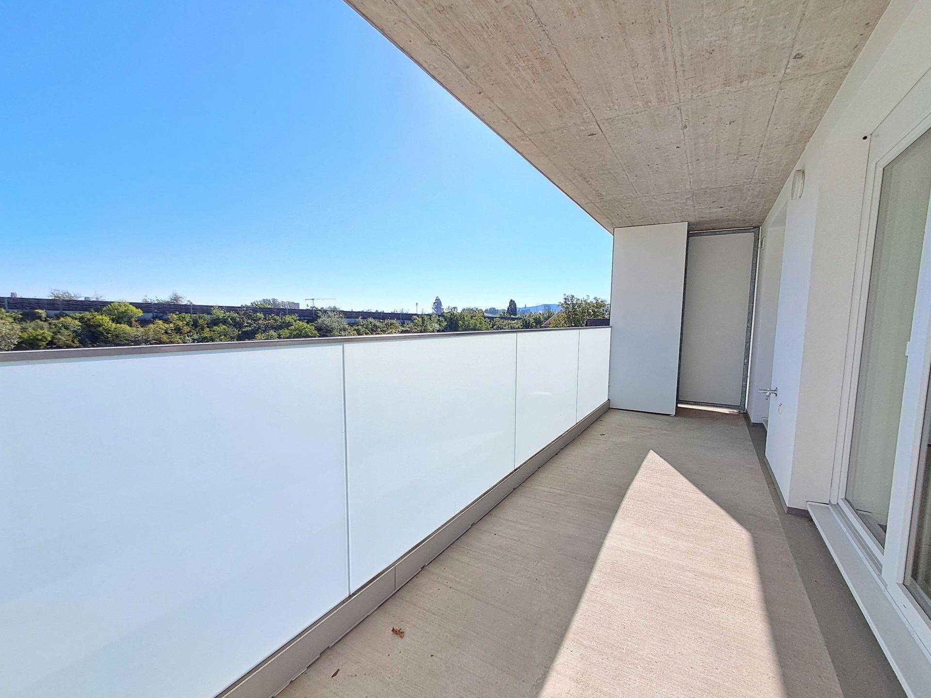 Heller Balkon mit moderner Glasbrüstung und weitem Blick ins Grüne unter strahlend blauem Himmel.