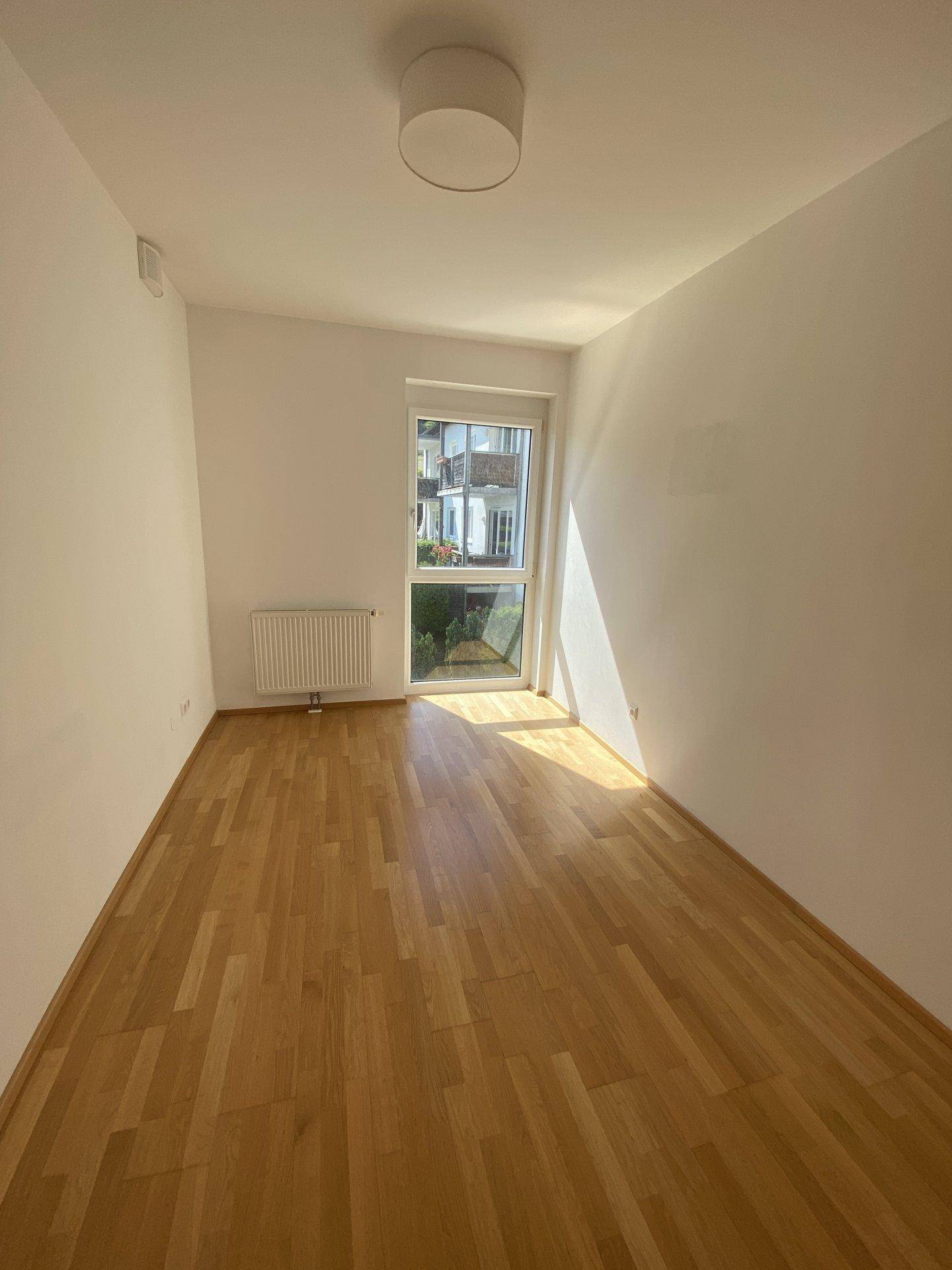 Helles Schlafzimmer mit Parkettboden und großem Fenster, das viel Tageslicht hereinlässt.