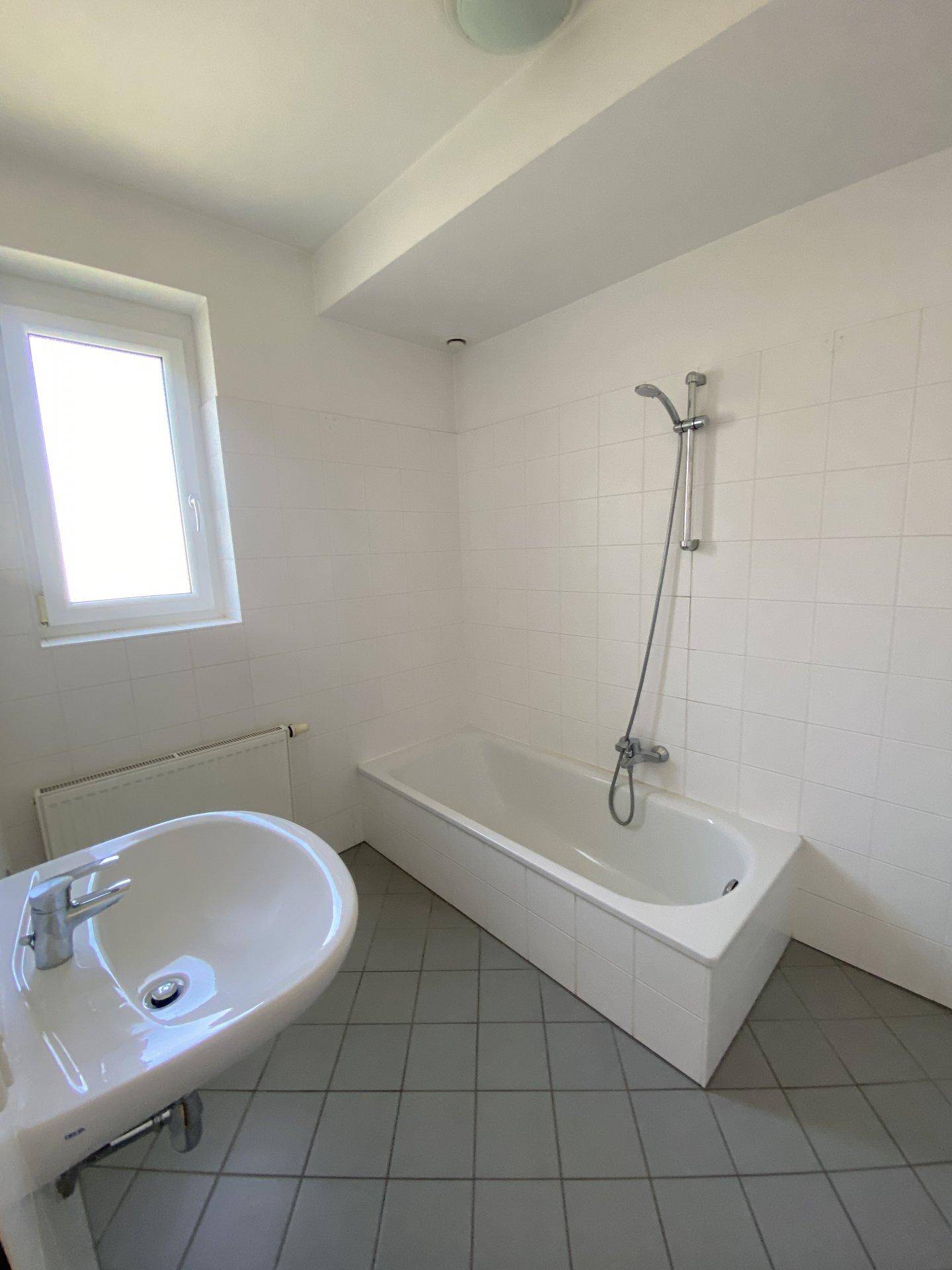 Helles Badezimmer mit Badewanne, Waschbecken, Fenster und weißen Fliesen.