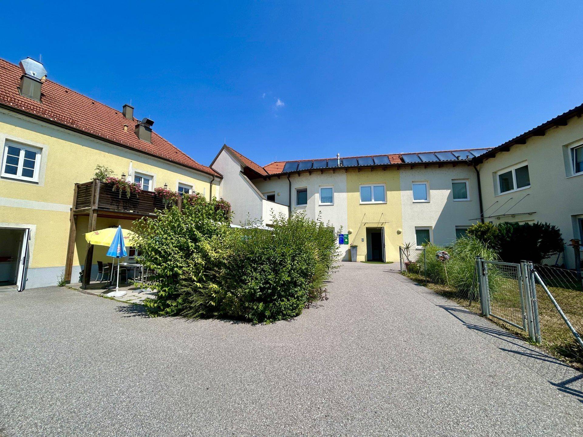 Ansicht des Gebäudekomplexes mit einem gepflasterten Bereich und Hecken unter strahlend blauem Himmel.