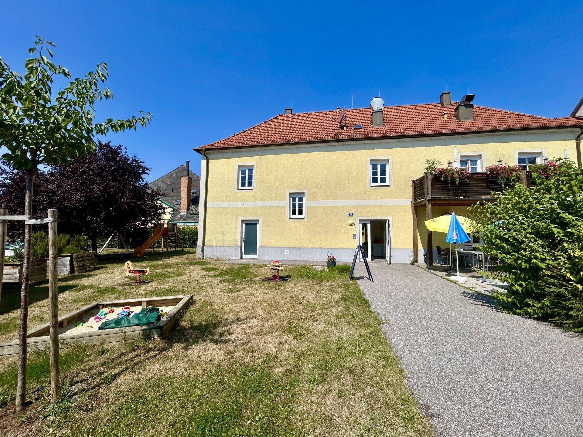 Gelbes Gebäude mit einem Gartenbereich, der einen Sandkasten und Spielgeräte für Kinder umfasst.