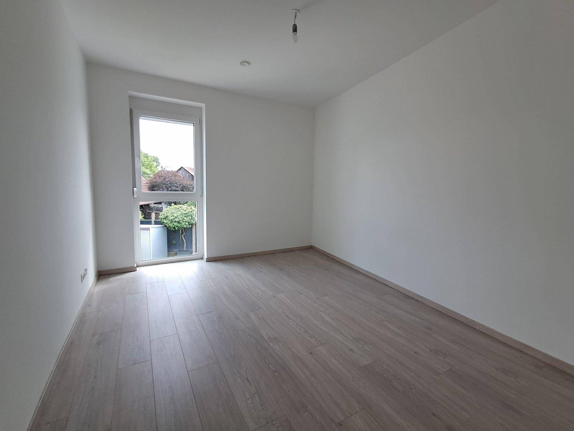 Freundliches Zimmer mit Laminatboden und Fenster mit Blick auf grüne Pflanzen.