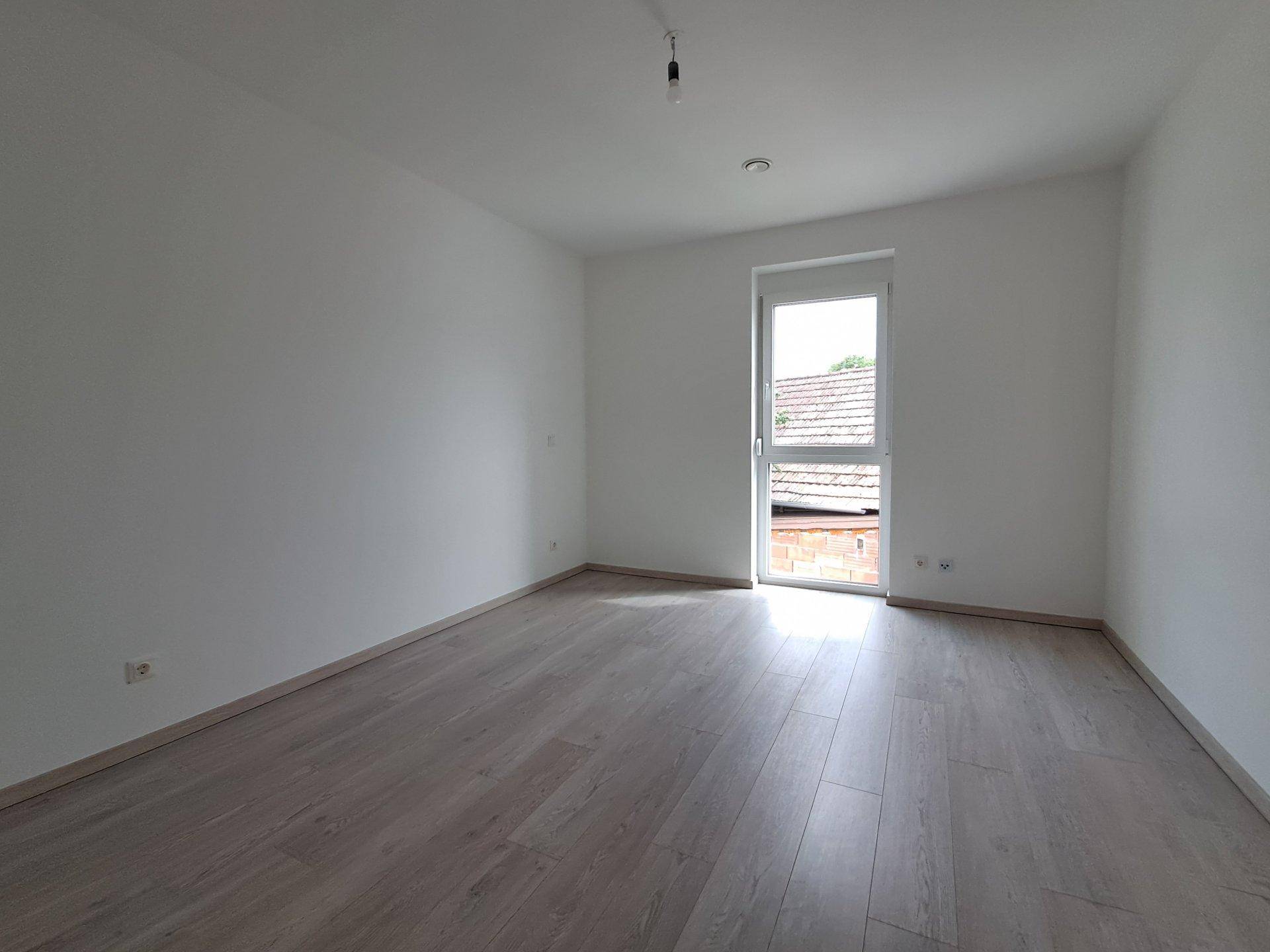 Leeres Zimmer mit Laminatboden und Fenster, das auf einen Außenbereich blickt.