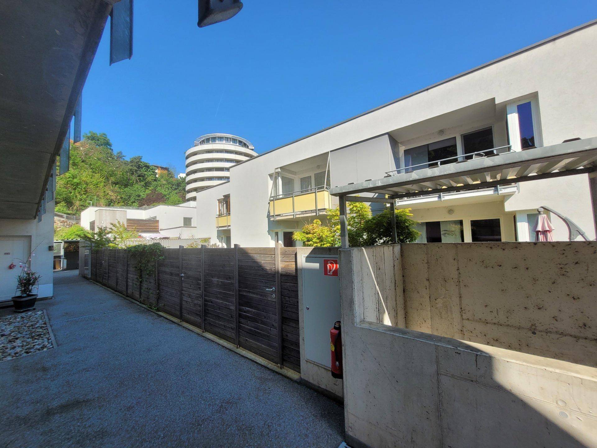 Weg zwischen Gebäuden mit Holzzaun und Blick auf Balkone sowie ein rundes Gebäude im Hintergrund.
