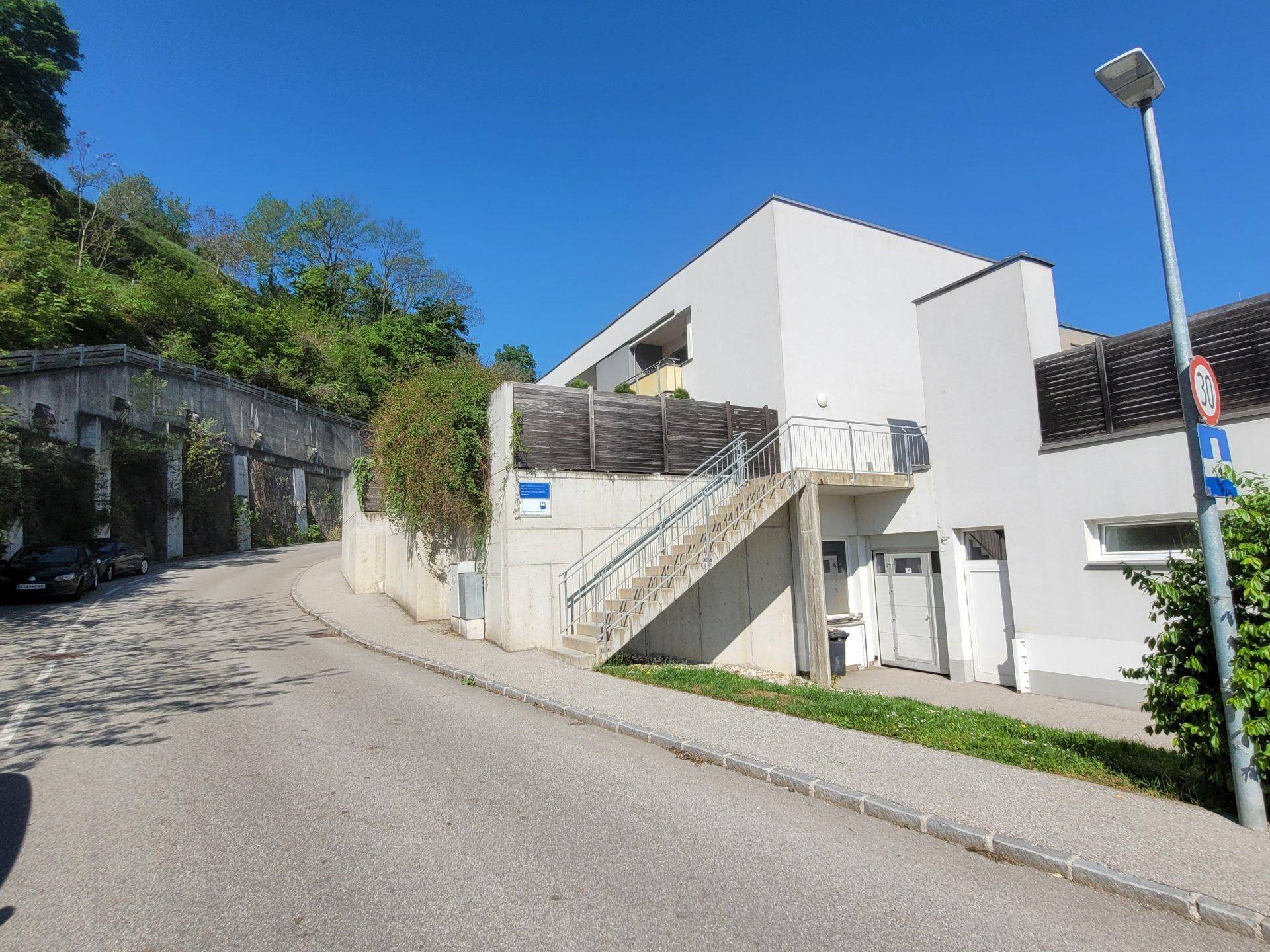 Weißes Gebäude an einem Hang mit Außentreppe und einer Straße im Vordergrund.