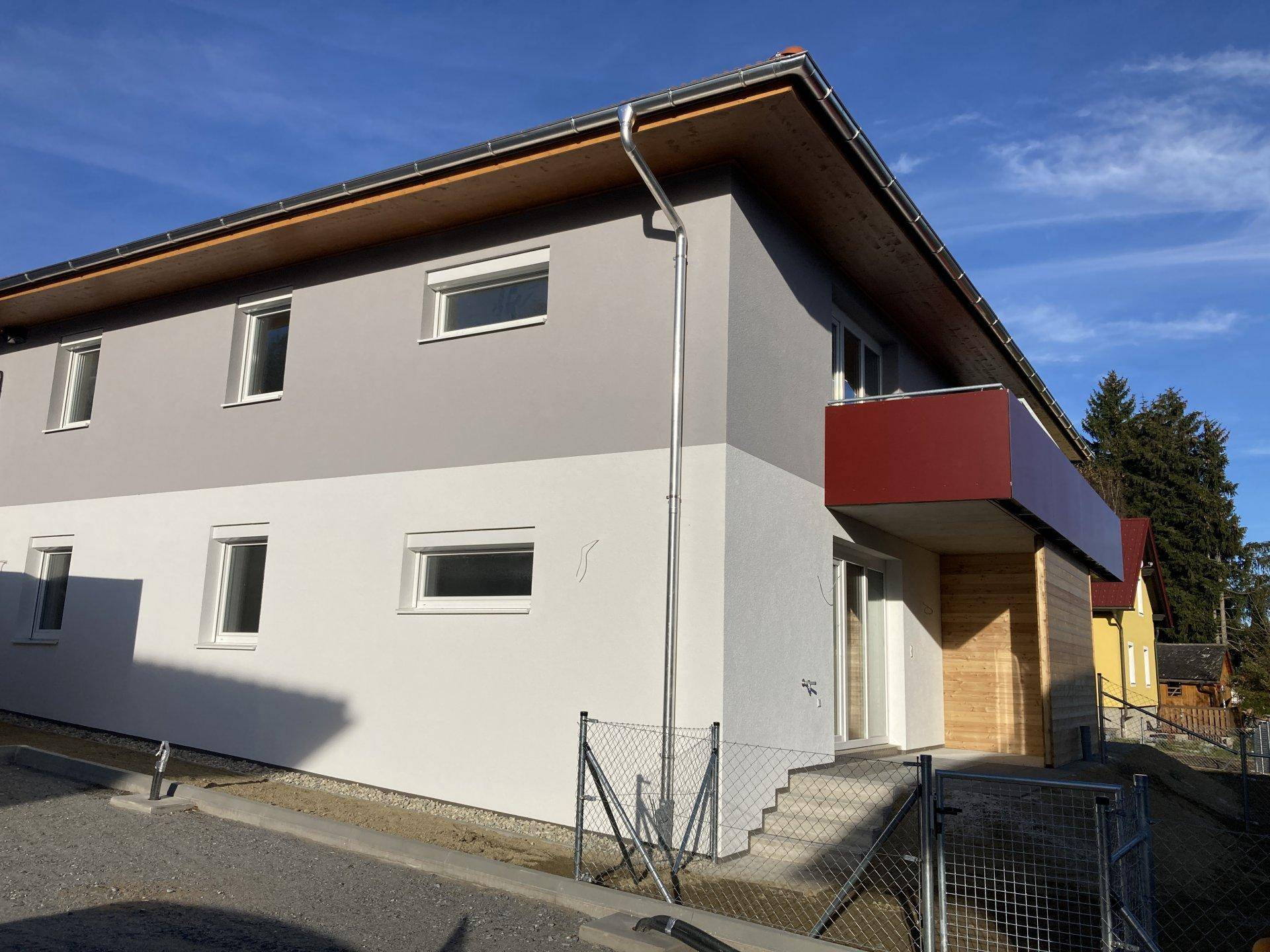 Seitenansicht eines modernen Neubaus mit grauer und weißer Fassade, rotem Balkon und Holzelementen.