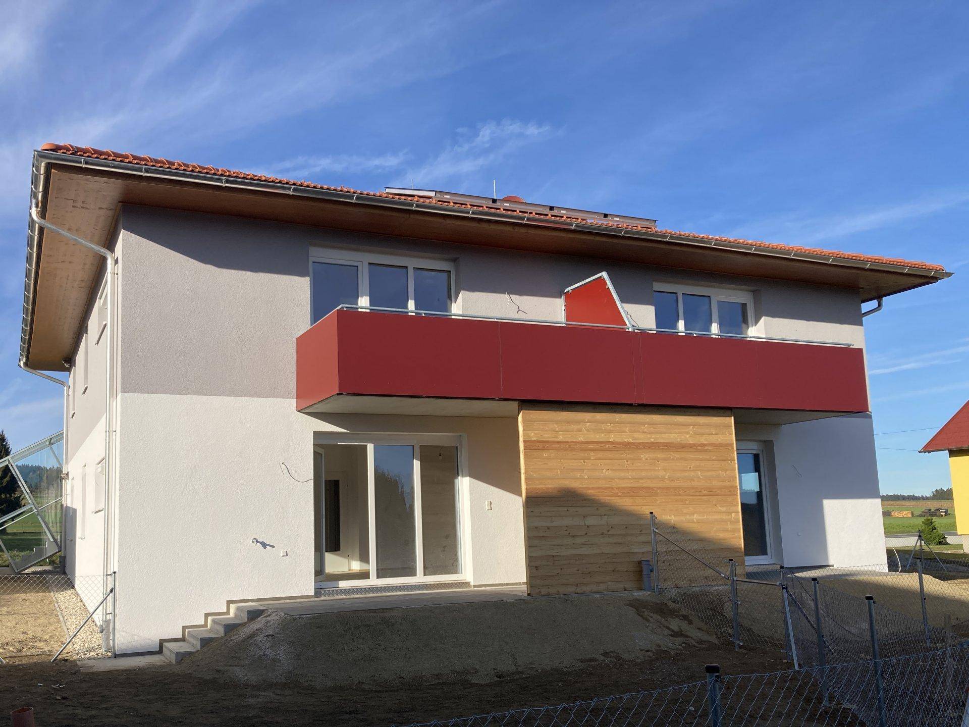 Vorderansicht eines Neubaus mit rotem Balkon, großen Glasschiebetüren und Holzverkleidung an der Fassade.