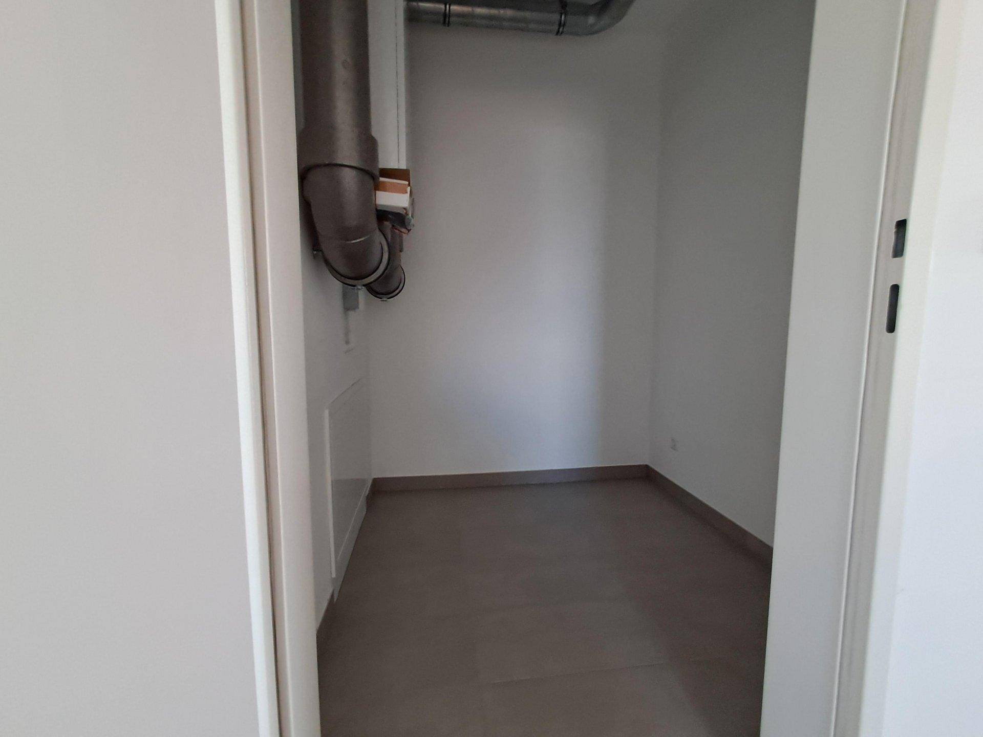 Kleiner Hauswirtschaftsraum mit grauen Fliesen, weißen Wänden und sichtbaren Rohrleitungen an der Decke.