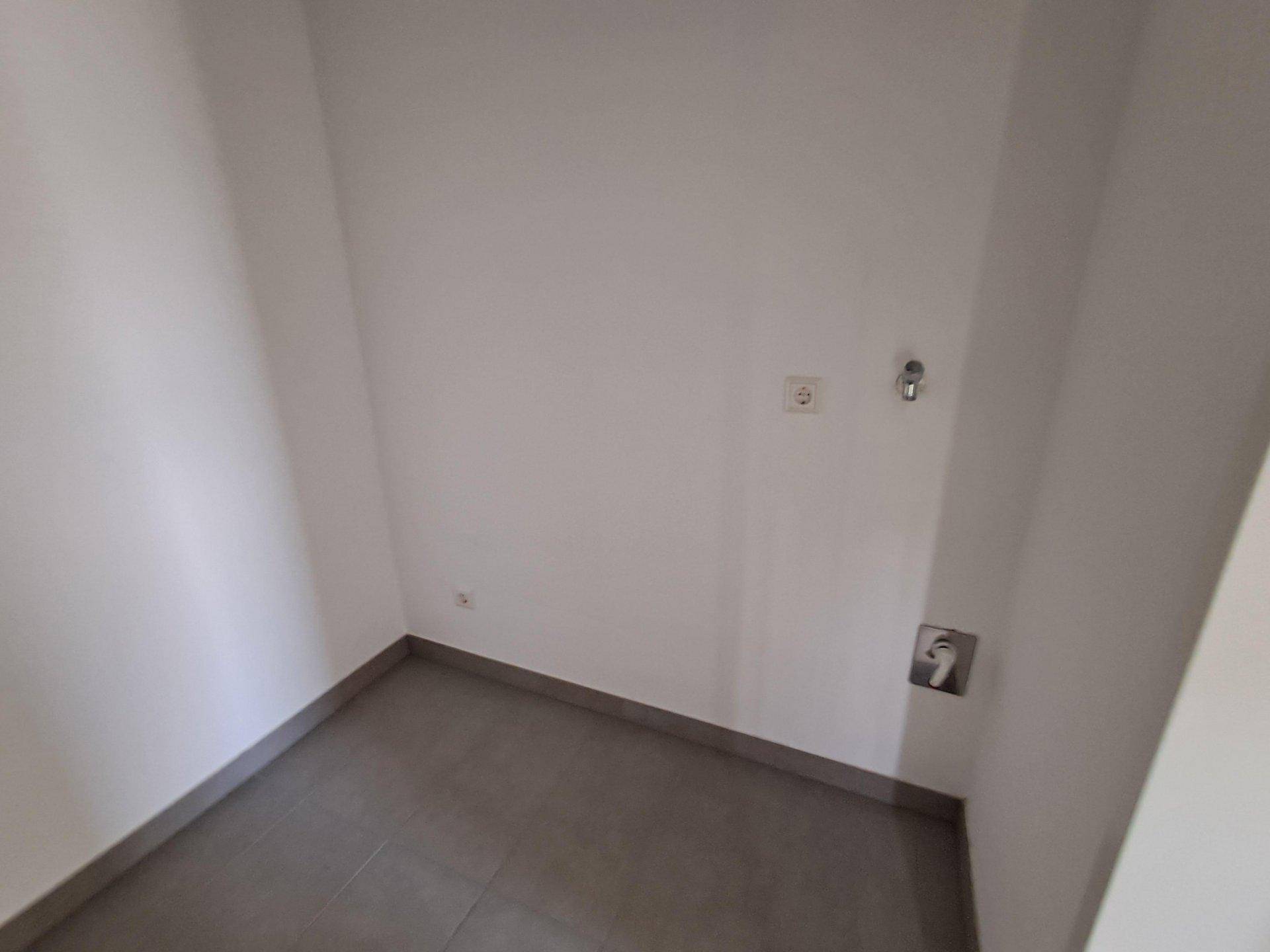 Kleiner Hauswirtschaftsraum mit grauen Fliesen, weißen Wänden und Anschlüssen für eine Waschmaschine.